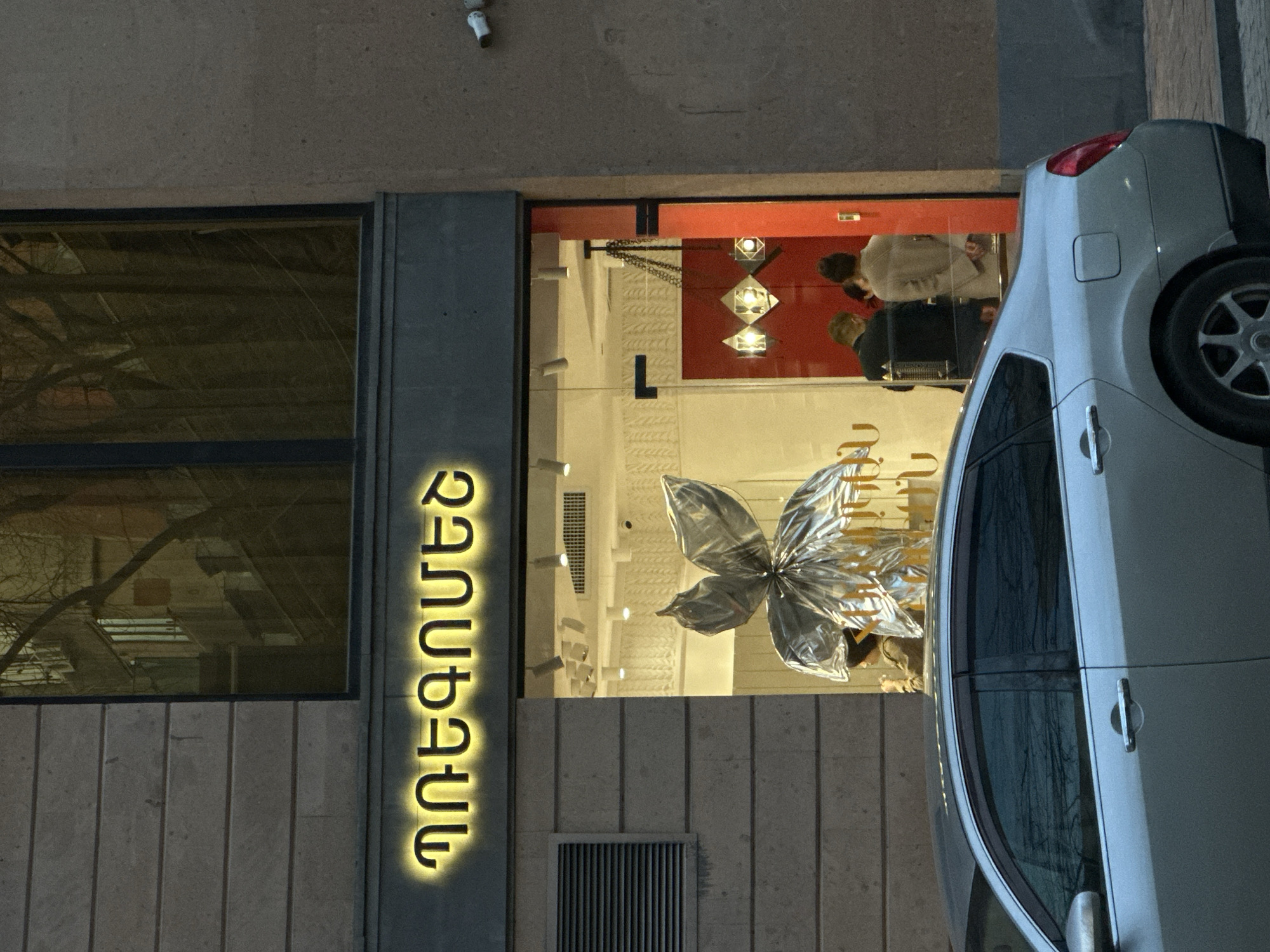 The image shows the storefront of a building in Yerevan, Armenia. The building has a sign with Armenian text illuminated in gold. Through the window, there's a silver decorative butterfly. Two people are visible inside the store, one wearing a light-colored coat. A silver car is parked on the right side of the image, partially blocking the view of the storefront. The building is constructed with light-colored stone or concrete.