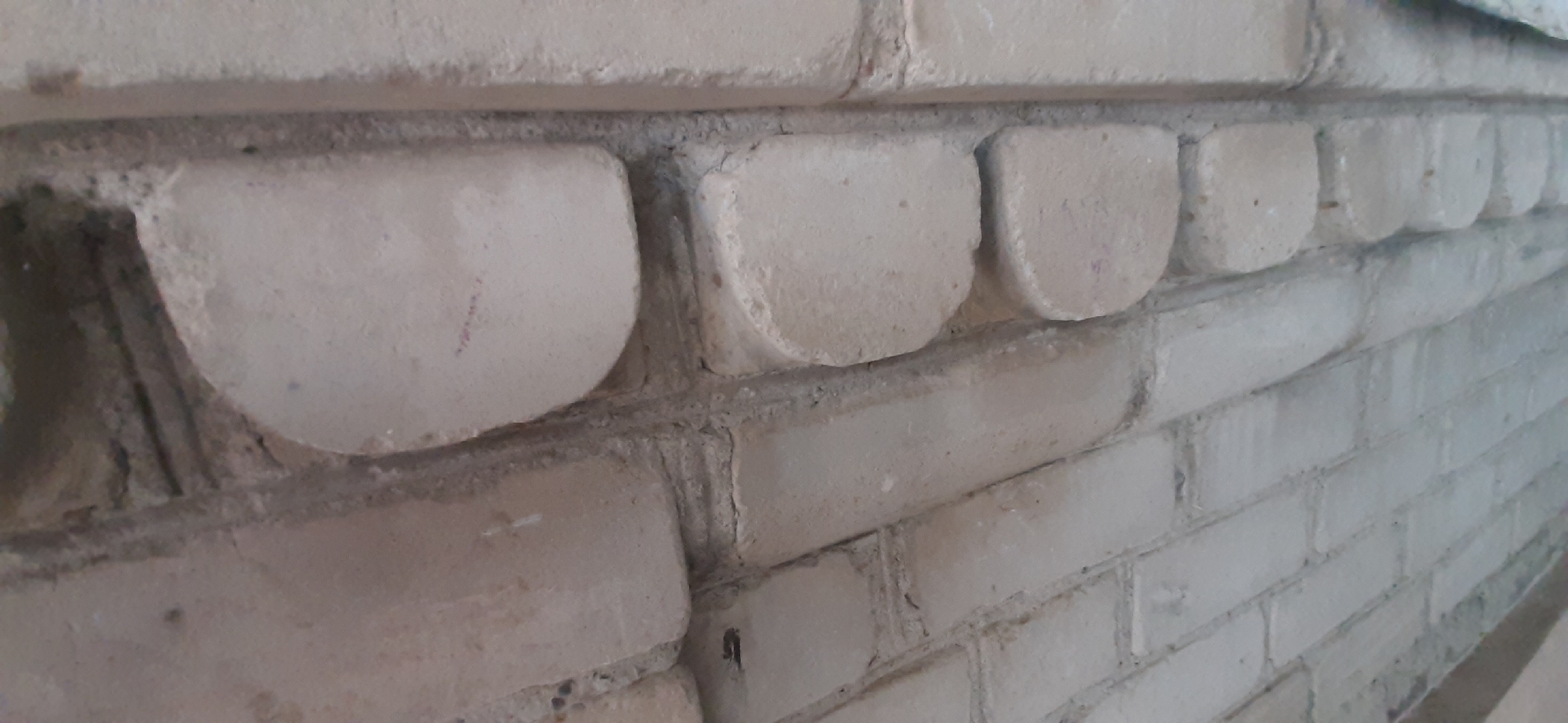 The image shows a close-up, angled view of a white brick wall with a decorative band of rounded brickwork along the top edge of the wall. The bricks appear to be weathered, with some slight discoloration and minor imperfections. The rounded bricks are evenly spaced and project slightly from the wall. The rest of the wall is constructed with standard rectangular bricks, also in white. The texture of the mortar between the bricks is visible, appearing somewhat rough and aged. There is no human presence or activity depicted in the image. The lighting suggests it might be daytime, but the specific time of day and weather conditions are not discernible. The scene is likely an exterior or interior architectural element in Yessentukskaya, Russia, given the provided location context, possibly a fireplace, a decorative border on a building facade, or a garden wall. No text is visible in the image.