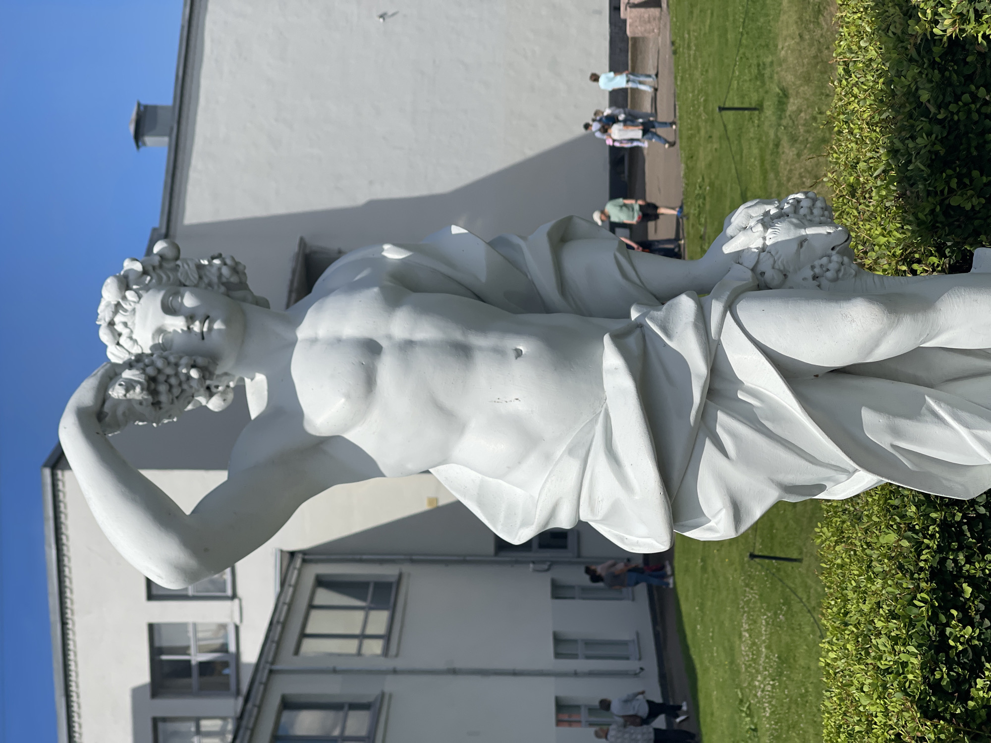 A white marble sculpture of a male figure adorned with grapes stands in front of a white building under a clear blue sky. The figure is muscular and holds a bunch of grapes above his head with his right arm, while his left arm is bent, holding another bunch of grapes and a lamb's head, which is a motif associated with Dionysus or Bacchus. The sculpture is draped in flowing fabric that accentuates its form. In the background, several people are walking on a green lawn, and the building appears to be a museum or cultural institution. The sculpture is located in the Vyborg Castle complex, Vyborg, Russia. The Vyborg Castle is a medieval castle located in Vyborg, Russia, on the Karelian Isthmus. It is a UNESCO World Heritage Site and is one of the most popular tourist attractions in the region. The castle was built in the 13th century by the Swedes and has been the site of many battles throughout history.