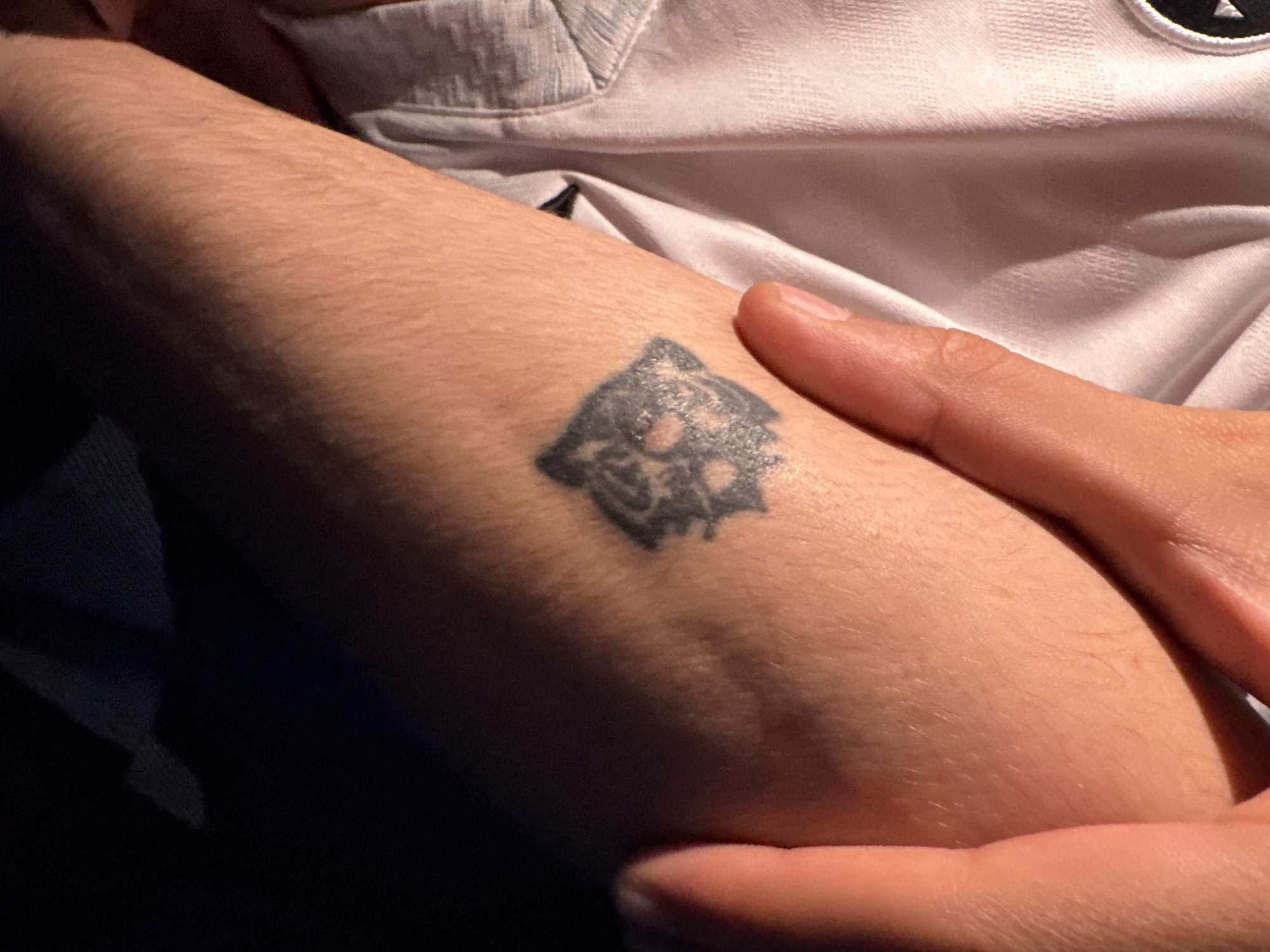 A close-up shot depicts a light-skinned arm with a dark tattoo located on the forearm. The tattoo appears to be a stylized image. A hand with short fingernails is also present, resting on the arm. The background shows a light-colored shirt and dark material of some kind. There is no information that can determine the location in Mexico City, Mexico.