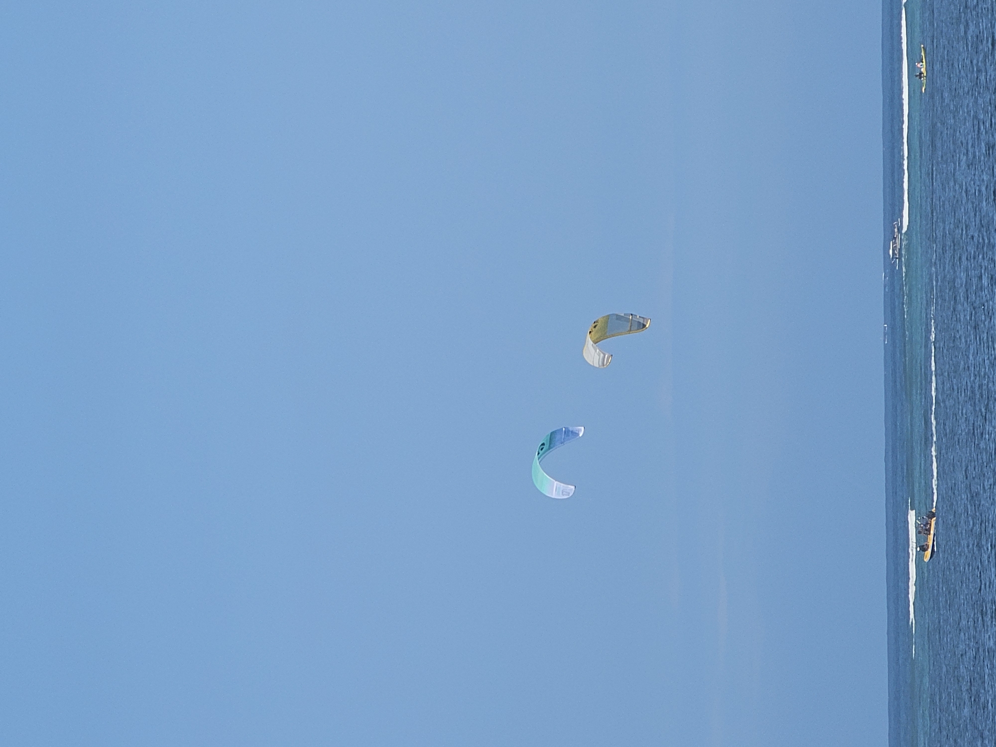 Two kitesurfing kites, one teal and white, the other yellow and white, are visible against a clear blue sky. Below the kites, the ocean is visible, with waves breaking in the distance. Several boats are on the water, including a yellow inflatable boat with people on board, and a longer boat further in the distance. The scene appears to be a coastal area. This view is consistent with many beaches in the Denpasar area of Indonesia, such as Kuta Beach or Seminyak Beach, which are popular for water sports.