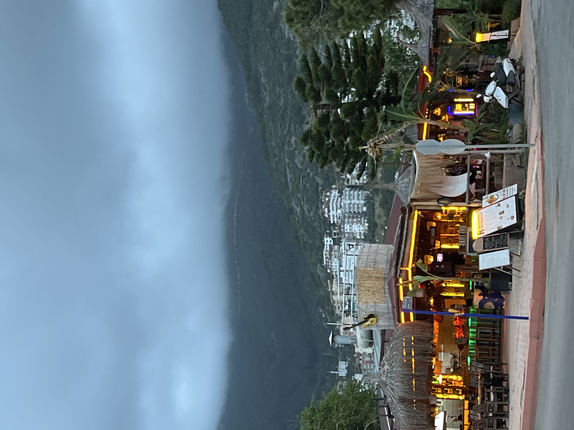 The image depicts a street scene in Alanya, Turkey, on a day with heavy cloud cover. On the right side of the frame, a road leads past a collection of beachside restaurants and bars, many with thatched roofs and illuminated with warm-toned lighting. Tables and chairs suggest an active outdoor dining area. Beyond these establishments, the cityscape of Alanya stretches along the lower slopes of a mountain range that fades into the cloudy sky on the left side of the image. The architecture of the city includes white buildings, indicating a Mediterranean style. The mountain is covered in greenery, with the upper portions obscured by the atmospheric haze. A guitar is visible on the side of a building, indicating the presence of music. A parked scooter and assorted signage are noticeable among the commercial establishments. The scene is generally static, with the focus on the environment and the ambiance of the restaurant area.