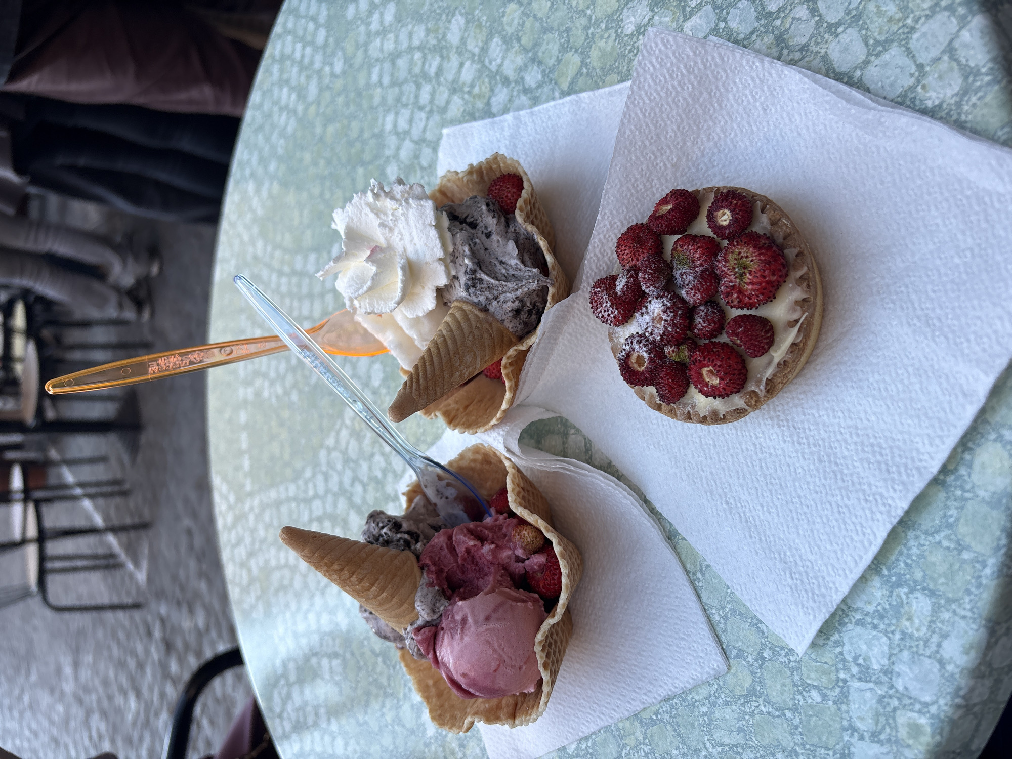 The image depicts a close-up view of two ice cream cones and a small strawberry tart on a table. The cones are waffle-style, and one is topped with whipped cream and dark ice cream, while the other contains a pink-hued ice cream and dark ice cream. Both cones have spoons inserted. The tart is round, with a pastry crust and filled with small strawberries dusted with powdered sugar. Each treat sits on its own white paper napkin. The table's surface has a pale green mottled pattern. The image likely was taken in Nemi, Italy, given the popularity of wild strawberries in this area. The environment suggests an outdoor café or restaurant setting.