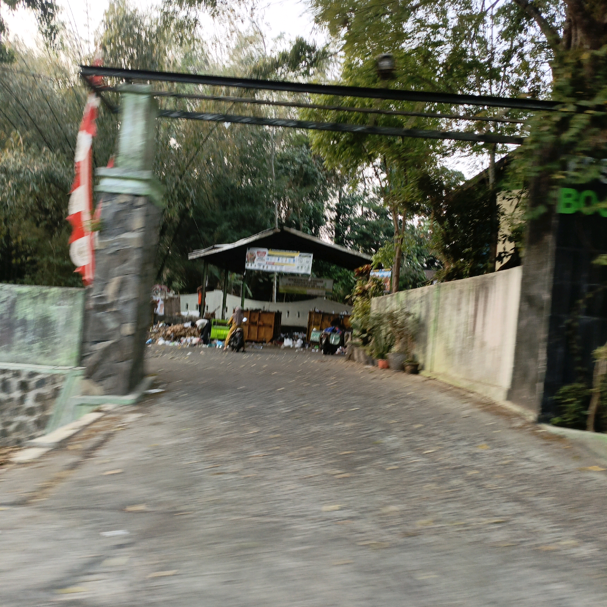 The image shows an outdoor scene of a waste sorting or collection facility located in Salatiga, Indonesia. The entrance to the facility is marked by a stone pillar with a red and white flag attached. A paved road leads into the facility, which features a covered structure with a pitched roof where waste bins are placed. Several people are visible, some interacting with the waste, and others are on motorcycles. There is a significant amount of waste scattered around the bins. The environment is characterized by a mix of trees and vegetation, and a wall runs along one side of the road. Green signage with text is visible on a wall to the right of the entrance. The overall impression is of a functional, somewhat untidy, waste management site.
