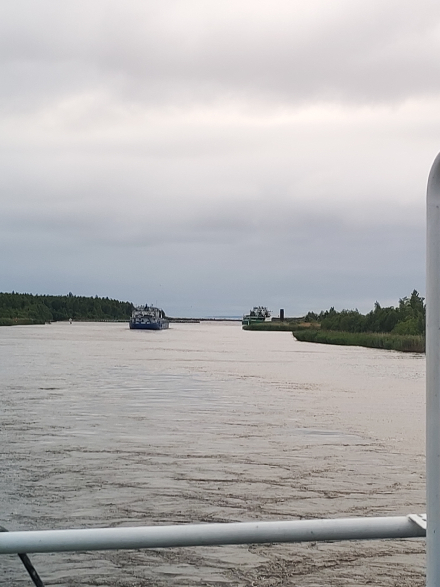 A wide, gray river flows through a rural landscape under an overcast sky. The riverbanks are lined with dense green trees and reeds. In the middle distance, two ships are visible. One ship, on the left side of the frame, is blue and white and appears to be a cargo vessel. The other ship, on the right, is green and white and seems to be a tugboat or a smaller utility vessel. The water is choppy, with visible ripples and currents. The foreground of the image is occupied by a white railing, suggesting the photo was taken from a boat or a dock. The overall atmosphere is calm and serene. The location is Ozernoe Ust'e, Russia, a port city on the northern tip of Lake Ladoga.
