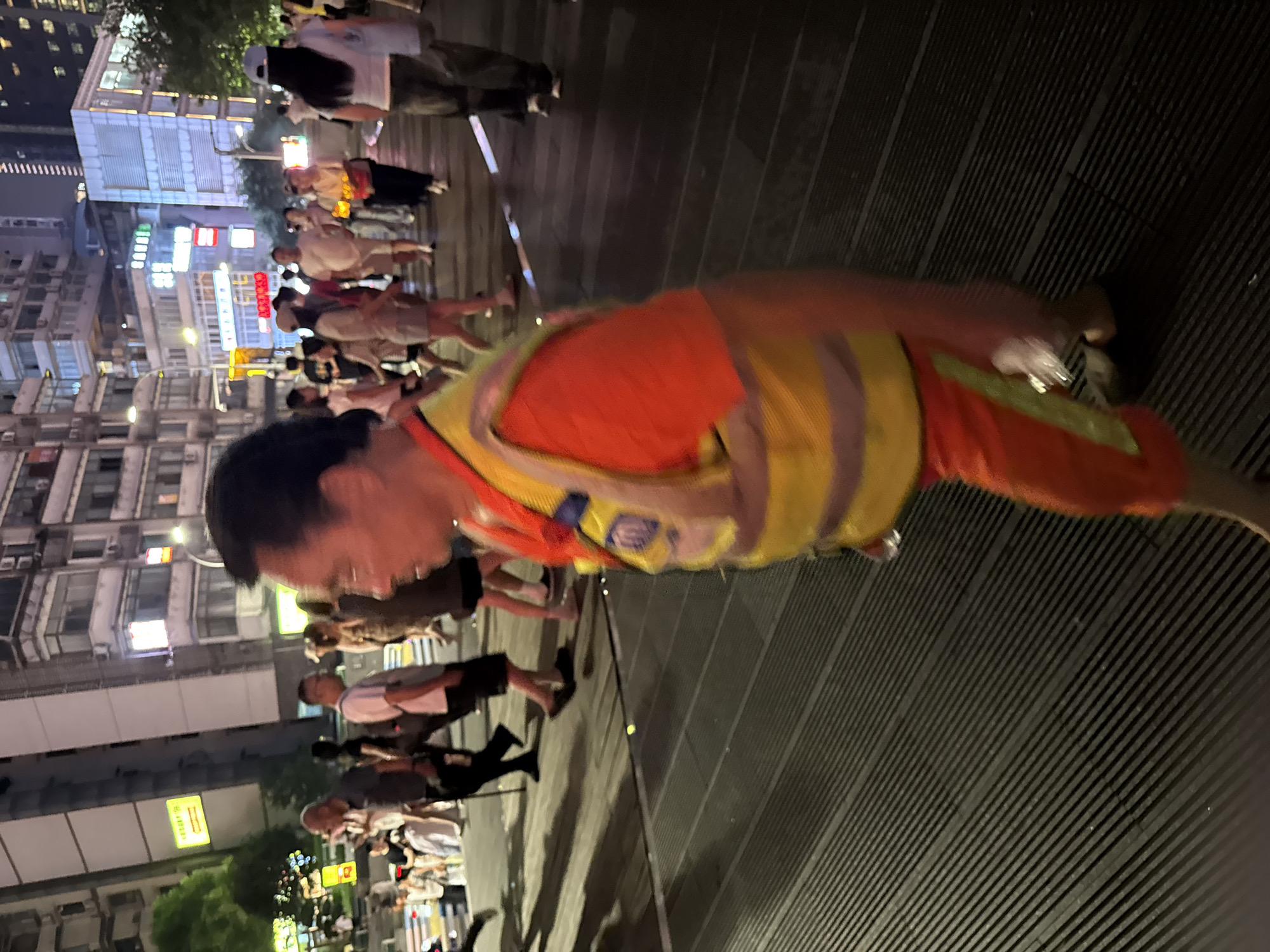 A man wearing an orange shirt and a yellow reflective vest is walking on a street in Yuzhong District, China at night. He has dark hair and is facing to the right of the frame. In the background, there are several buildings, some of which are illuminated. There are also other people walking in the background, as well as streetlights and signs. The ground is a dark, ribbed surface.