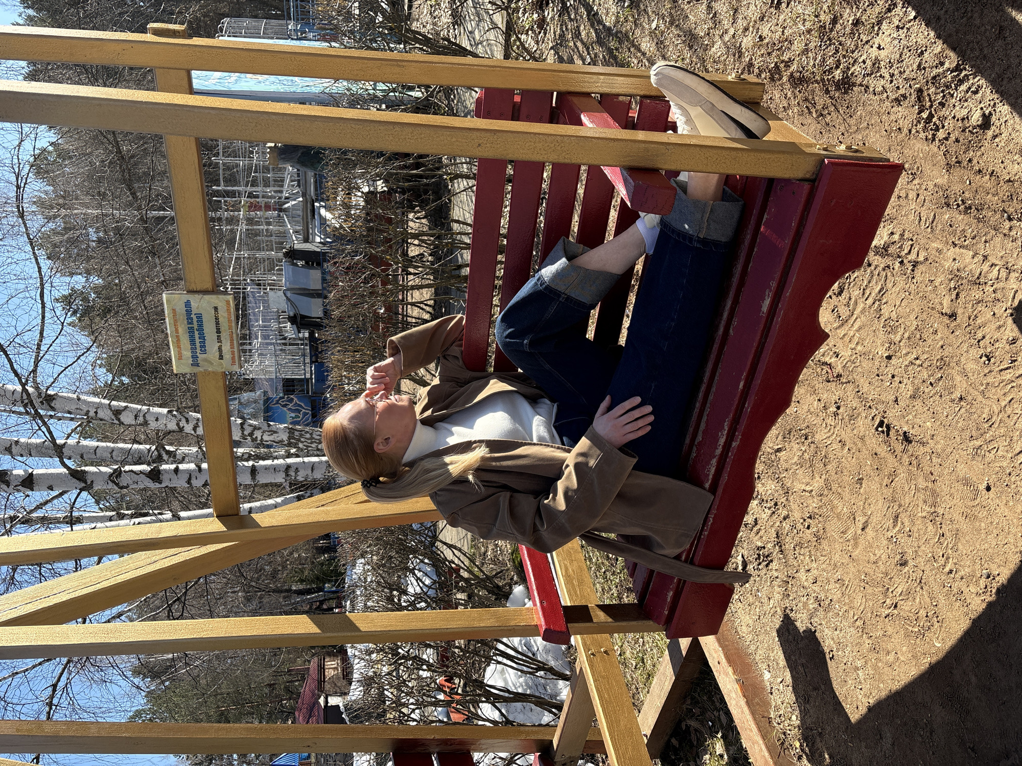 The image captures a relaxed scene featuring a woman lying on a rustic wooden swing-bench in an outdoor setting, likely a park or recreational area in Chaikovsky, Russia. The swing-bench itself is constructed with golden-brown wooden beams supporting a red-painted slatted seat.
The main subject, a woman with light reddish-brown hair tied back in a low ponytail, is dressed in casual, warm attire suitable for a cool, sunny day. She wears a white turtleneck top, a brown jacket, and dark blue jeans with rolled-up cuffs, revealing white socks and light-colored sneakers. Her head is tilted back, and she appears to be looking upwards or into the distance, with one hand near her chin, suggesting a moment of thought or relaxation.
The setting is characterized by clear blue skies and bare trees, including distinctive white-barked birches, indicating early spring or late autumn. The ground beneath the swing is dry, sandy soil with visible footprints and shadows cast by the swing structure and the woman, suggesting bright, direct sunlight. In the background, beyond the trees, there are hints of structures or water, adding depth to the scene.
A light-colored sign is affixed to one of the swing's wooden beams, displaying text in Russian. The visible text reads:
* "Деревянная качель" (Wooden swing)
* "(свадебная)" (wedding)
* "Надпись для фотосессий" (Inscription for photoshoots)
* "Только для фотосессий" (Only for photoshoots)
This indicates the swing is a decorative prop specifically intended for photography. The bright sunlight, clear weather, and the woman's tranquil posture evoke a peaceful and contemplative mood.