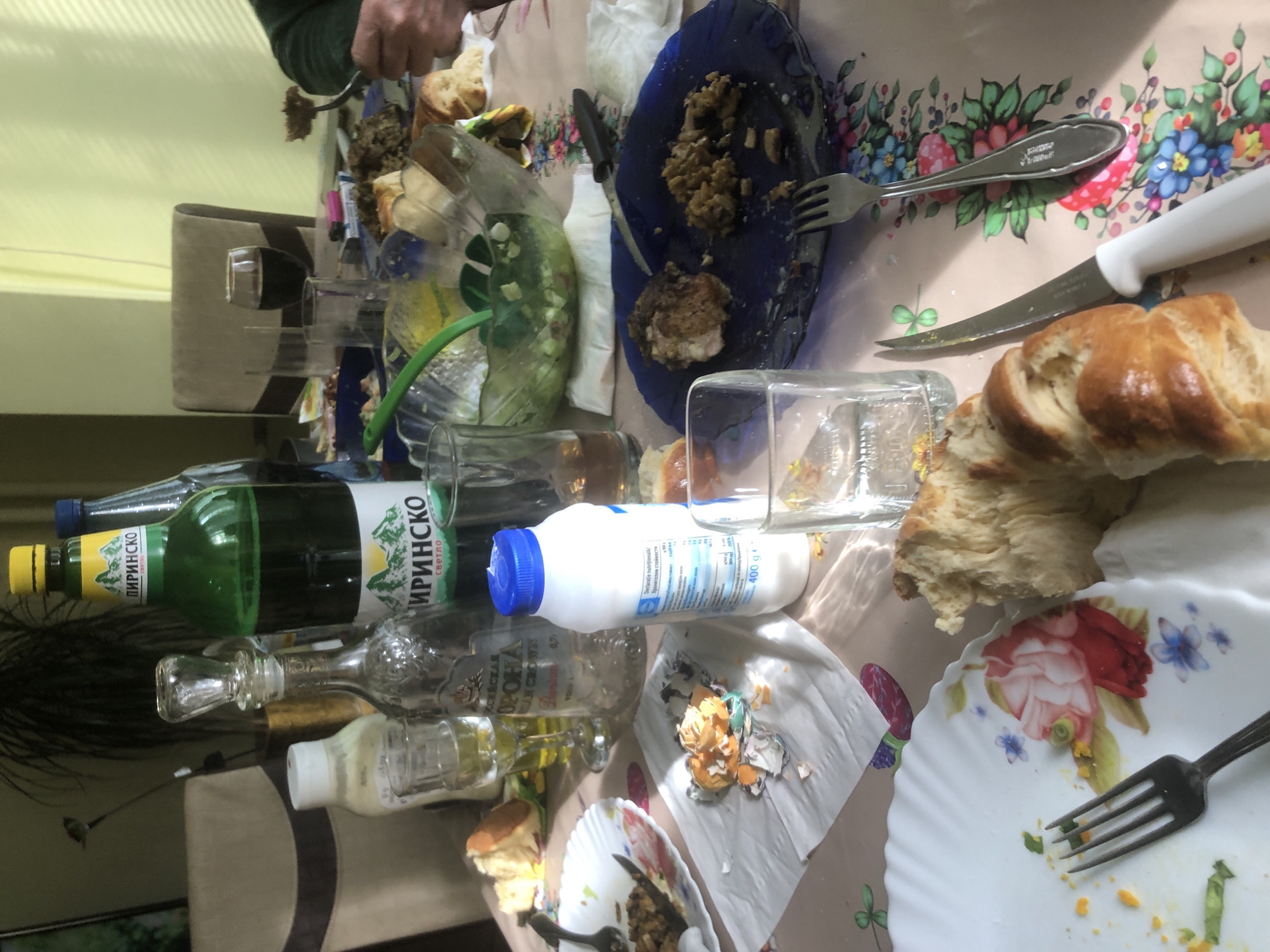 The image shows a table covered in a floral-patterned tablecloth with remnants of a meal. Food items include bread, unidentified cooked food on plates, and eggshells on a napkin. Several bottles are present, including "Пиринско" (a Bulgarian beer brand), an empty vodka bottle labeled "РОССИЙСКАЯ", and a bottle of what appears to be mayonnaise. Also present are a knife, forks, and glasses. A hand holding a fork is visible at the top of the frame. The scene is located in Bulgaria based on the bottles and brand names visible. There is no location information that would indicate the city of Sofia.