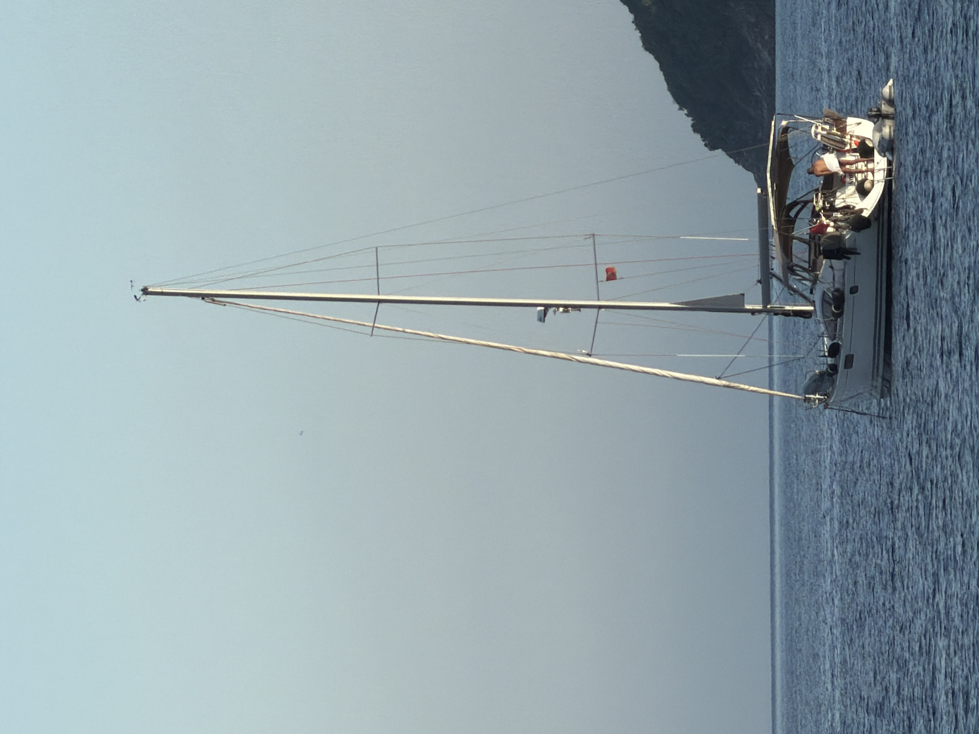 A sailboat is anchored in calm, blue water. The mast is tall and thin, reaching towards a clear, light blue sky. A red flag hangs from the mast, further down from the top. Several people are visible on the deck of the boat. A dark, green, mountainous landmass is in the background. The location is suggested to be Budva, Montenegro.