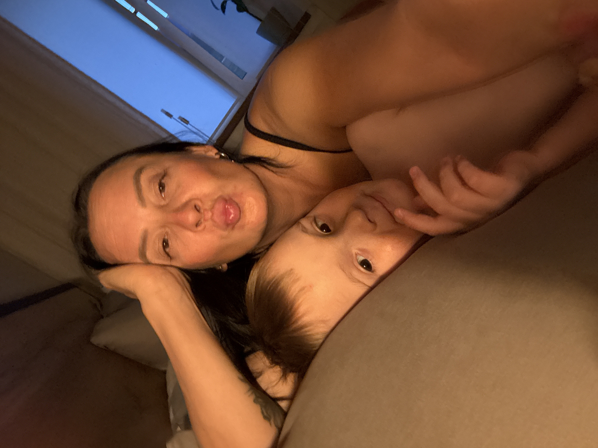 The image appears to depict a selfie of a woman and a young child lying down. The woman, with dark hair, is positioned in the foreground, making a playful face with her tongue slightly protruding. The child, with lighter hair, is nestled against her, gazing towards the camera. They both seem to be lying on a bed or similar soft surface, with a window visible in the background, suggesting an indoor setting. The image seems to have been taken indoors, but there is no specific indication of it being in Chaikovsky, Russia.