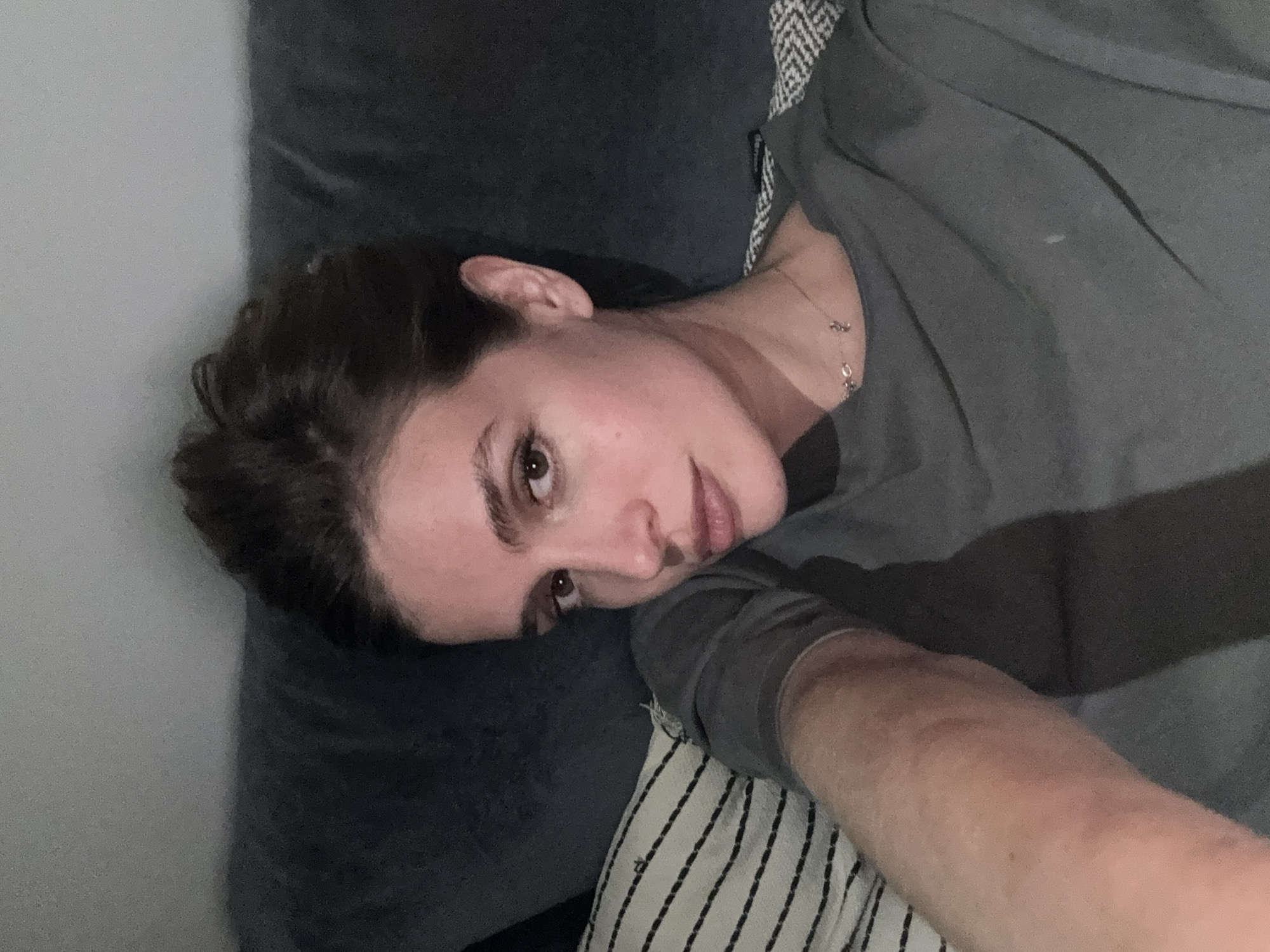 A woman is lying on a gray couch with a black-and-white striped pillow. She is wearing a gray shirt and has a necklace. Her hair is brown. The background is a plain white wall. The location is not determinable from the image.