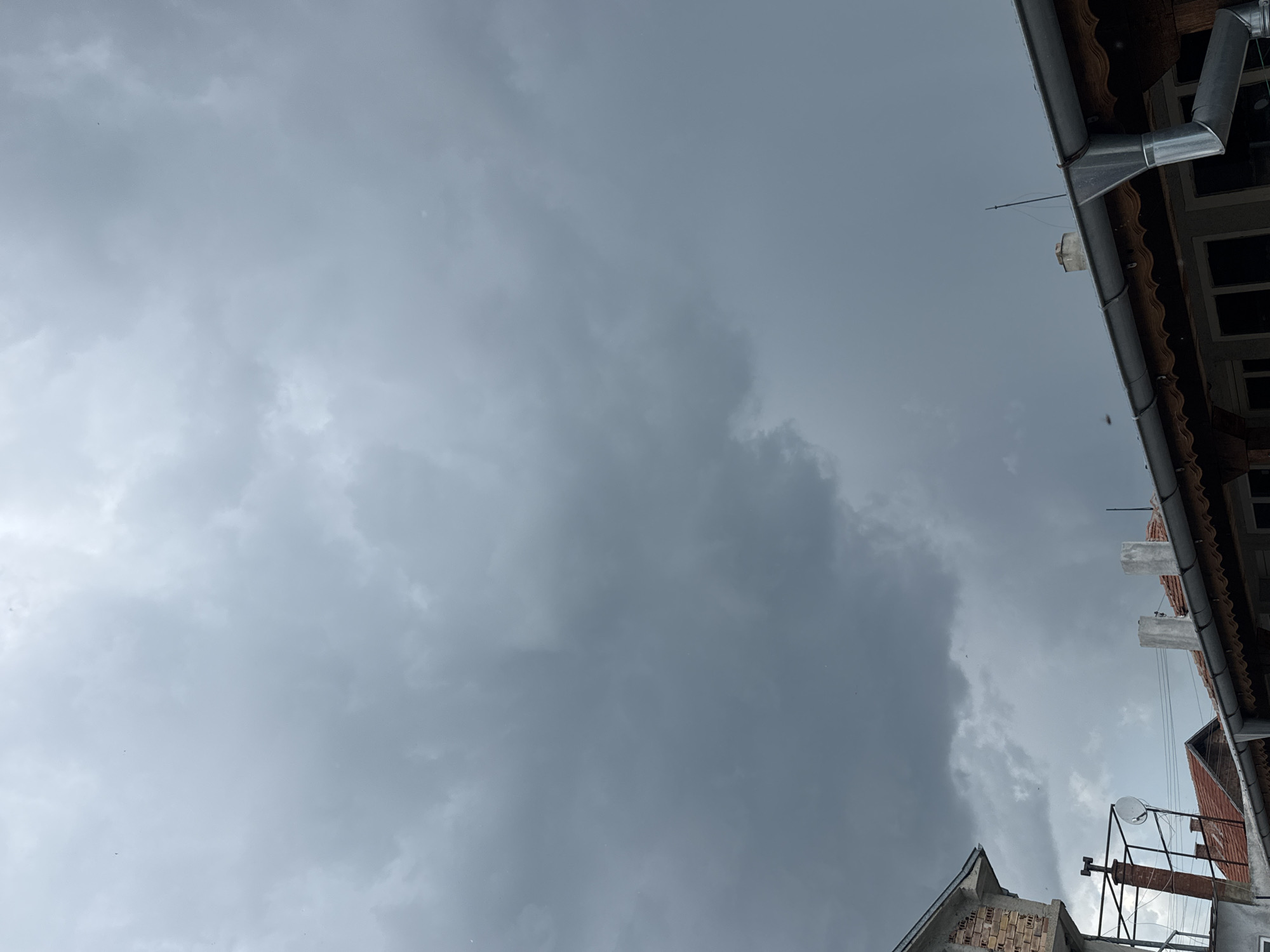 The image shows a view looking upward towards a cloudy, overcast sky. The bottom of the image shows sections of buildings with brickwork, roofing, satellite dishes, and other architectural elements. In the upper-right, there is a building's guttering and a small window, indicating a close proximity to structures. The overall composition is tilted. The location is Sarnevo, Bulgaria.