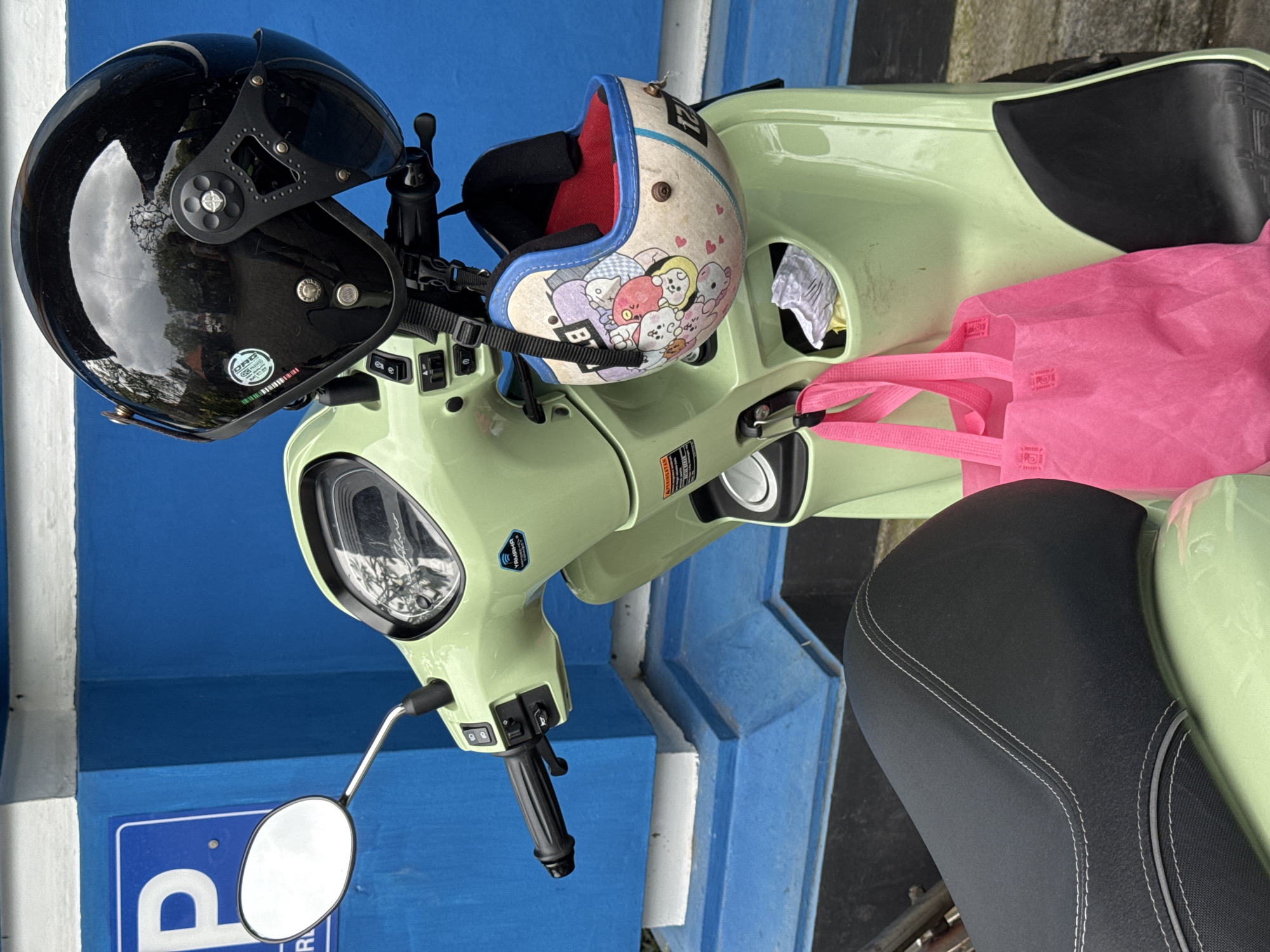 A light green scooter is parked outdoors, viewed from a slightly high angle against a vibrant blue wall. The scooter features a sleek, modern design with a black seat adorned with white stitching.
Two helmets are positioned on the scooter. A glossy black open-face helmet with a short visor rests on the handlebars. Its reflective surface clearly shows reflections of trees and a bright sky, suggesting a sunny daytime. A white sticker on this helmet displays "DBN," "ECE R22.05," and "SNI," indicating compliance with European and Indonesian safety standards. Also hanging from the handlebars is a cream-colored, open-face helmet, worn and decorated with an array of pastel-colored cartoon characters and small heart motifs, trimmed with blue piping.
A bright pink fabric bag is placed on the scooter's floorboard or front area, adding a pop of color to the scene. The scooter's handlebar area is visible, showing controls, a rearview mirror, and a "Yamaha FAZZIO" decal, identifying the make and model. Below the mirror, a blue sign partially visible on the wall appears to be a parking sign, with the top of a 'P' symbol indicating it might be for disabled parking.
The setting, with its bright colors and casual arrangement of personal items, suggests a typical street scene in Kuta, Indonesia, where scooters are a common mode of transport. The items indicate the scooter is either ready for use or has just been used, hinting at daily activities.