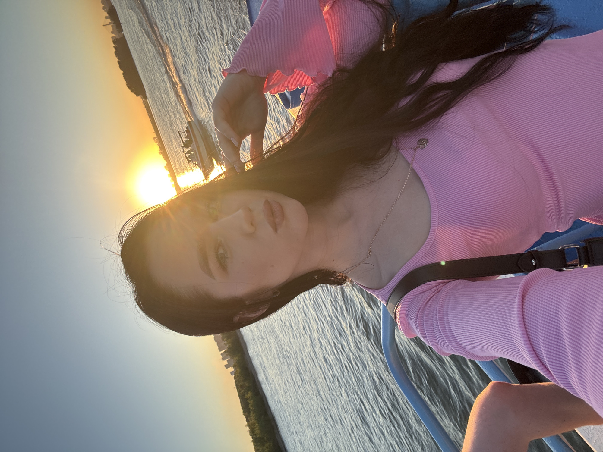 A woman with long, dark hair, wearing a pink ribbed top and a necklace, takes a selfie on a boat at sunset. The sun is setting over a body of water, casting a warm glow on the scene. In the background, a cityscape and trees are visible on the shore, with a speedboat in the distance. The image was taken on the Ob River in Novosibirsk, Russia.