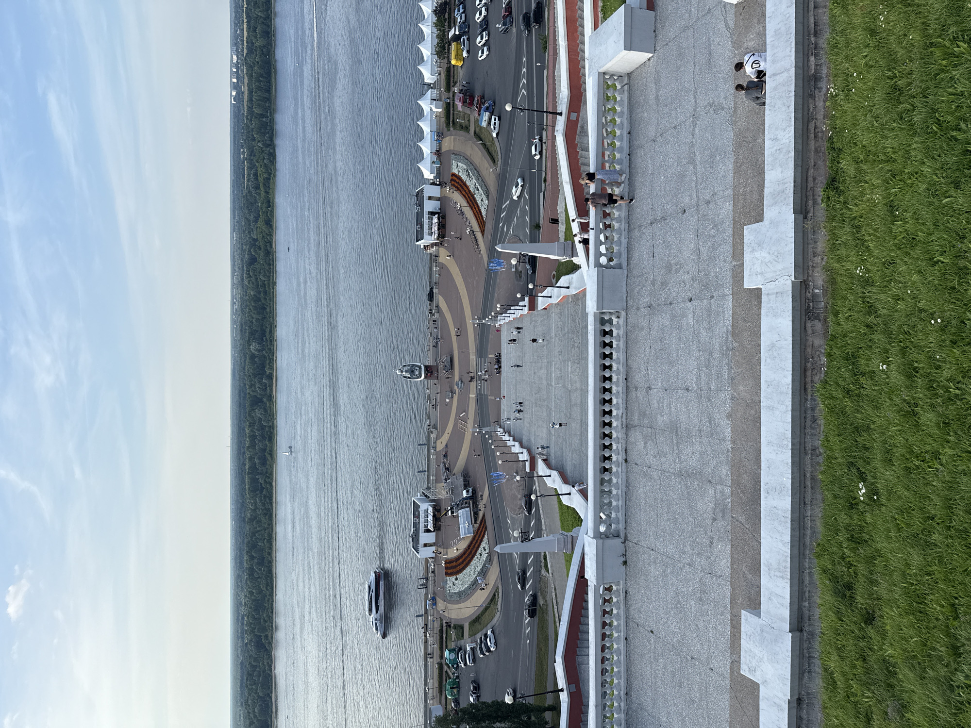 This aerial view captures a bustling riverfront promenade in Nizhny Novgorod, Russia. The scene is dominated by a wide, calm river on the left, reflecting the pale blue sky with wispy clouds, suggesting a pleasant daytime with fair weather. A line of green trees marks the opposite bank of the river.
The promenade itself is a central focus, paved with textured concrete and featuring a decorative circular area with flowerbeds. A road runs parallel to the promenade, with several cars, including a yellow truck and a white minivan, parked or in motion. Pedestrian activity is evident, with scattered individuals walking along the promenade and near the road. Two boats are visible on the river, one larger and modern, the other smaller and white. White tents are set up along the edge of the road, hinting at a possible event or market.
On the right side of the image, a manicured grassy area with scattered white wildflowers borders the promenade. A white concrete barrier with decorative railing separates the promenade from the grass. Several people are visible standing on and near the barrier, some appearing to be conversing, while others are walking. The overall atmosphere seems relaxed and lively, characteristic of a summer day in a popular public space. The time of day appears to be late afternoon or early evening, given the soft light and the angle of the shadows.
