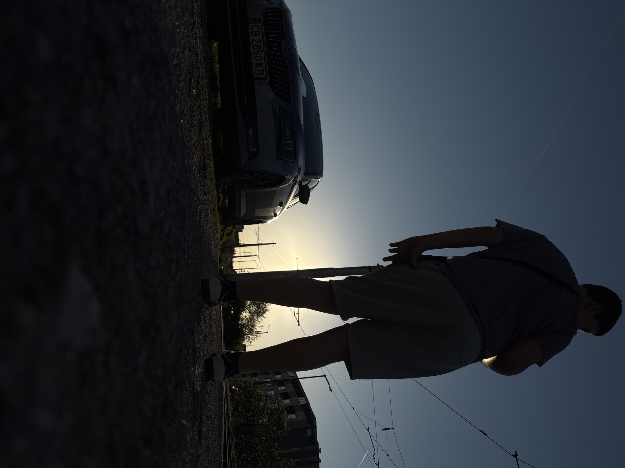 The image shows a man standing near a gray car with the license plate "CB 2850 KB". The man is wearing shorts, a t-shirt, and sneakers. The scene is silhouetted against the setting sun. There are tram lines and power lines visible, and the location appears to be in an urban area. Based on the license plate and architectural style of the building in the background, the city is most likely Sofia, Bulgaria.
