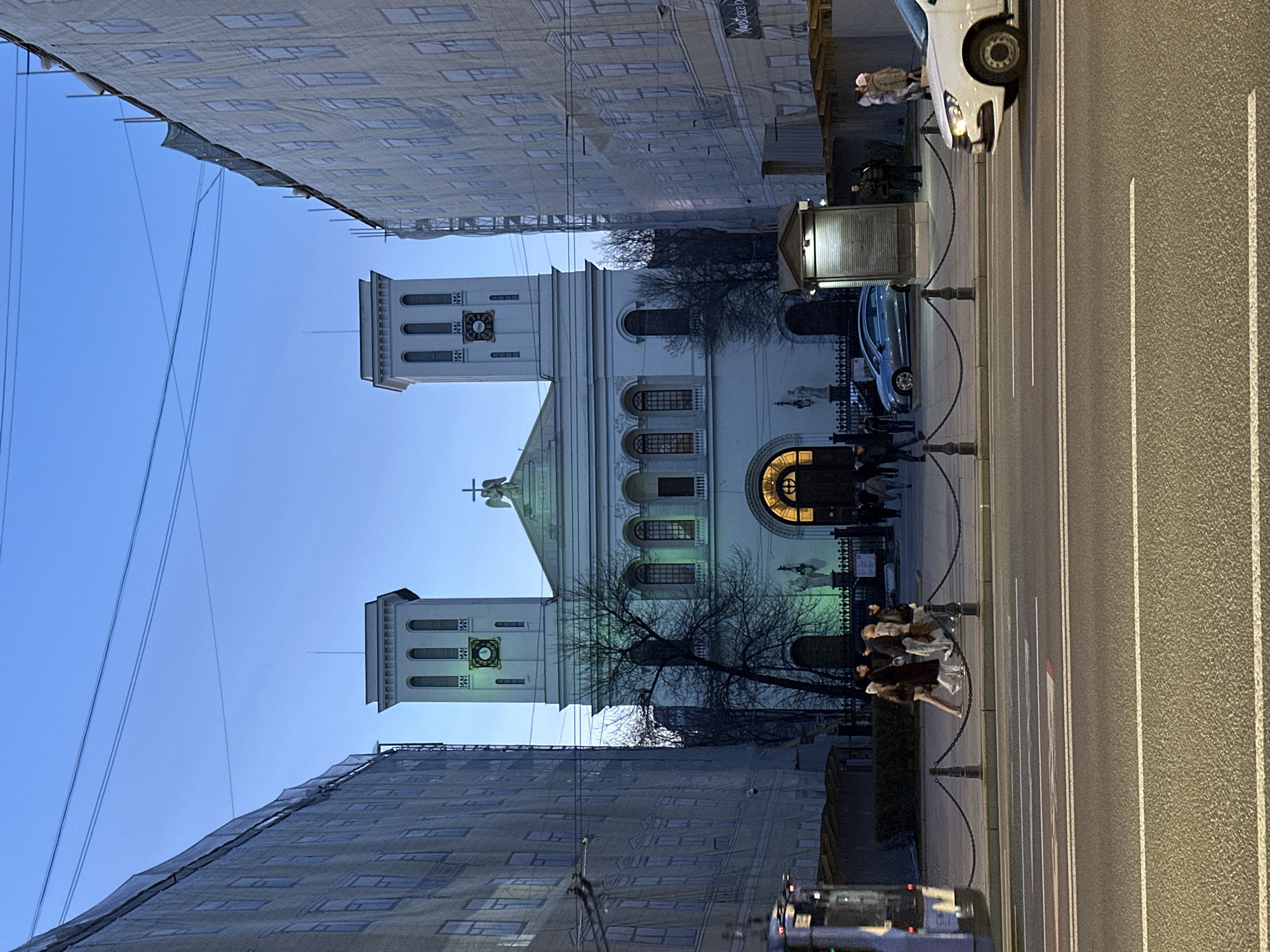The image depicts St. Anne's Church, also known as Annenkirche, in Saint Petersburg, Russia. The church is framed by buildings on either side, one of which is covered in a construction tarp. The church has two prominent towers and a central structure with arched windows and a round window above the entrance. People are gathered in front of the church and along the sidewalk, next to the road. Cars are seen parked along the street. The sky is clear blue.