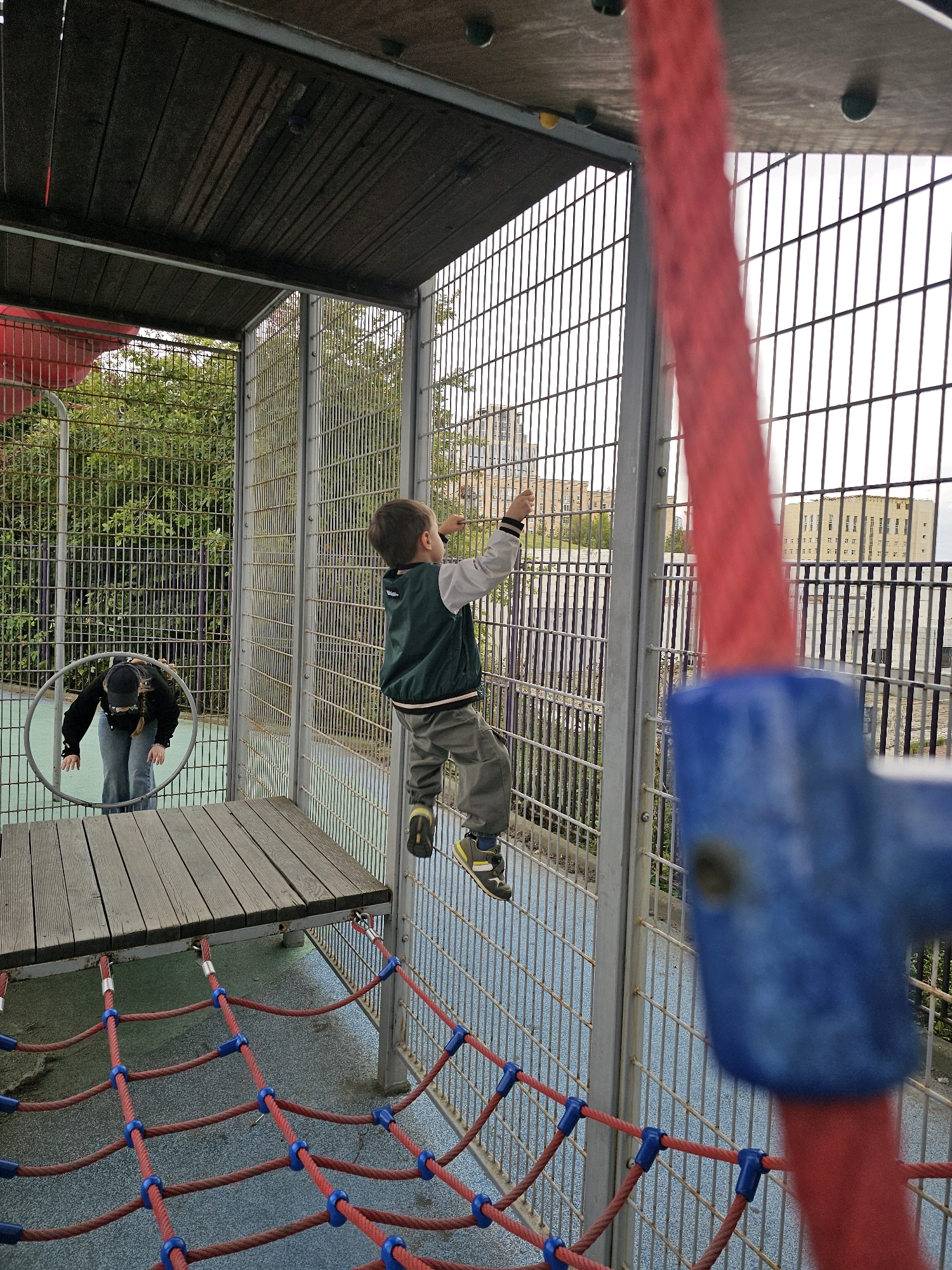 The image depicts a children's playground structure, likely in an urban setting, with two children present. In the foreground, a young boy with brown hair is actively climbing a metal grate structure, wearing a green vest over a gray long-sleeved shirt and gray pants. His right leg is extended upwards, and his left hand is reaching for the structure. To the left, a young person wearing a black hooded sweatshirt, black baseball cap, and blue jeans is bending over, appearing to interact with a circular metal hoop on a wooden platform. The playground features a wooden platform, metal fencing, and a red rope net with blue connectors. In the background, trees and multi-story buildings are visible, suggesting a densely populated area. The sky is overcast.
The location is identified as the Zaryadye Park children's playground, near the Moscow Kremlin, in Moscow, Russia.