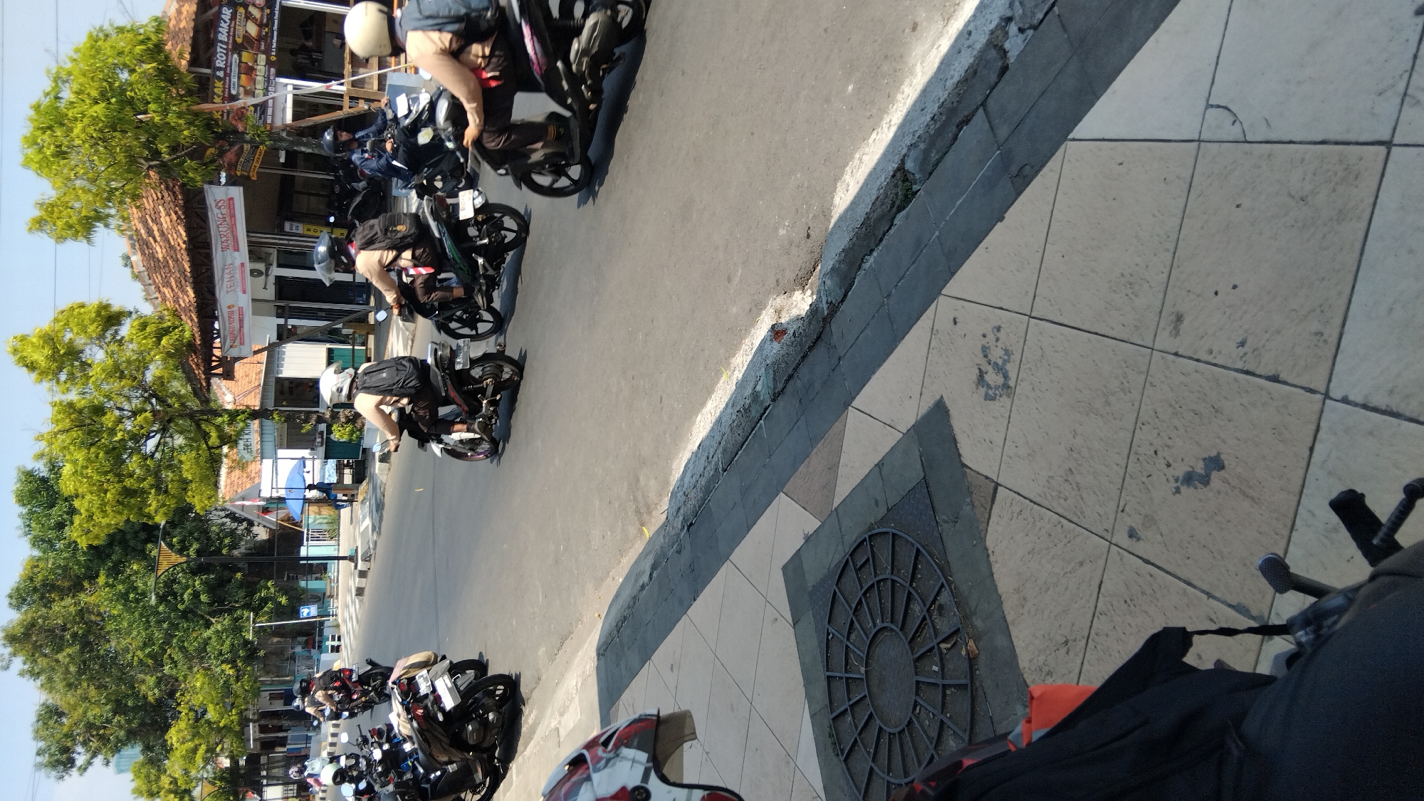 In Batang, Indonesia, several individuals on motorcycles are seen riding on a street. The scene appears to be in a commercial area, with shops lining the street, including one with signs for "TENAN WARUNG SS" and "KAKAK & ROTI BAKAR." The sky is clear and sunny, casting shadows on the road and sidewalk. Some of the motorcyclists are wearing helmets and backpacks. The foreground shows a sidewalk with patterned tiles and a manhole cover, and the edge of a black object, possibly a motorcycle seat or bag, is visible in the lower left corner.