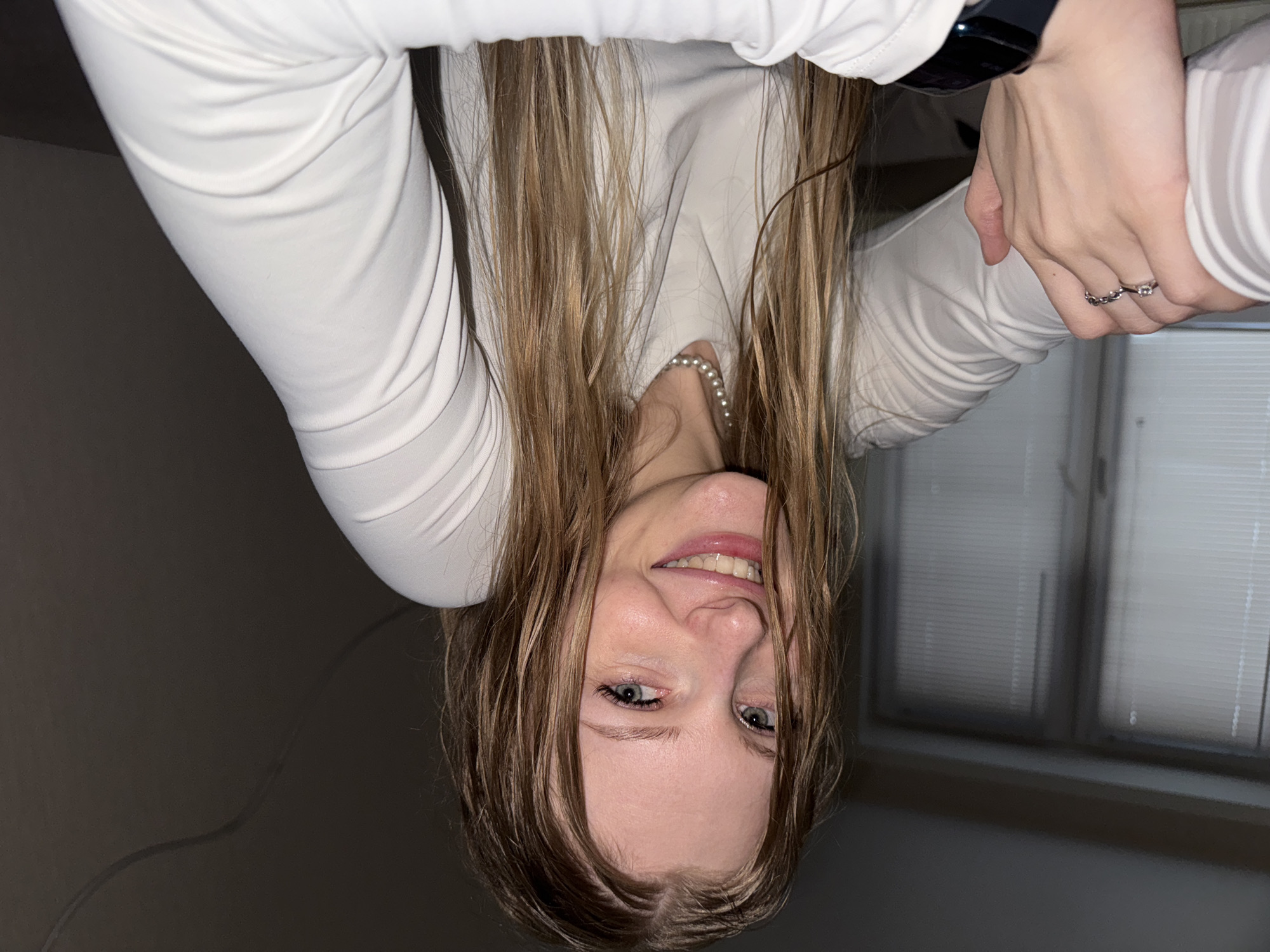 This media file features a young woman in an indoor setting, potentially a room in Perm, Russia. She is depicted upside down, looking directly at the camera with a happy, smiling expression that reveals her teeth. Her long, light brown hair, which appears to be damp or wet, falls around her face.
She is wearing a light-colored, long-sleeved top. Her accessories include a string of pearls around her neck, two rings on her right hand (one with a chain-like pattern and the other with a jewel), and a dark smartwatch on her left wrist.
The background is dimly lit. To her right, a window with horizontal blinds is partially visible. To her left, there's a dark, plain wall with a subtle cord or cable running across it. The low lighting suggests it might be evening or a dimly lit interior. The overall mood is casual and playful, captured through her inverted pose and cheerful demeanor. There is no visible text in the image.