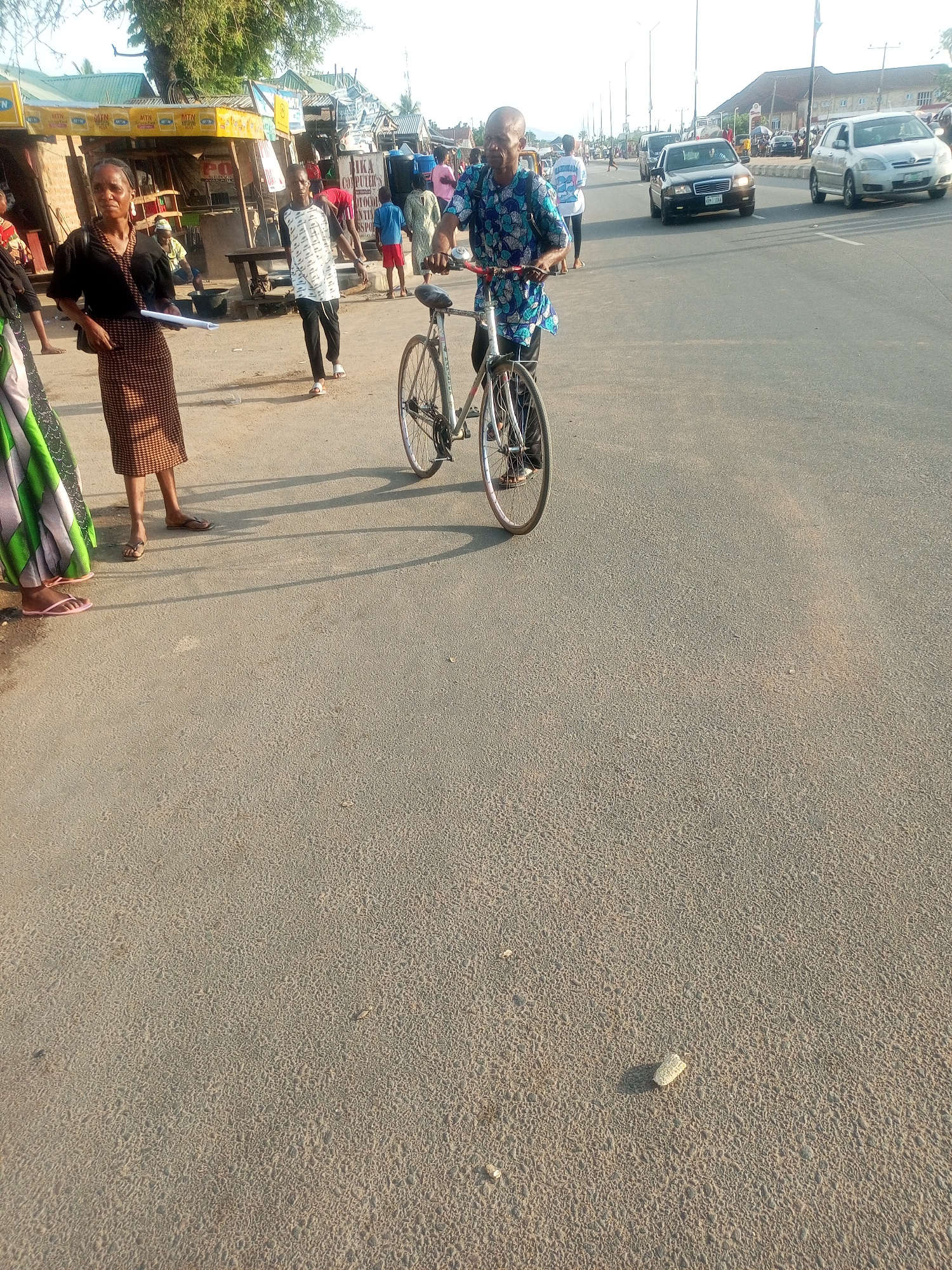 The image depicts a street scene in Jalingo, Nigeria. In the foreground, a man with a bald head wearing a blue and white patterned shirt rides a bicycle. Beside him, a woman in a black top and a patterned skirt stands with a rolled-up document in her hand, appearing to be engaged in conversation or waiting. Behind them, several other people are visible, some walking and others possibly standing near market stalls. The setting suggests a bustling outdoor environment with a road and what appear to be small shops or kiosks lining the side. In the background, cars are driving on the road, indicating a moderately busy street. The lighting suggests it is daytime, possibly late afternoon given the long shadows cast on the road. The overall atmosphere appears to be ordinary daily life unfolding in a public space. Text is visible on some of the market stalls, including "MTN" logos.