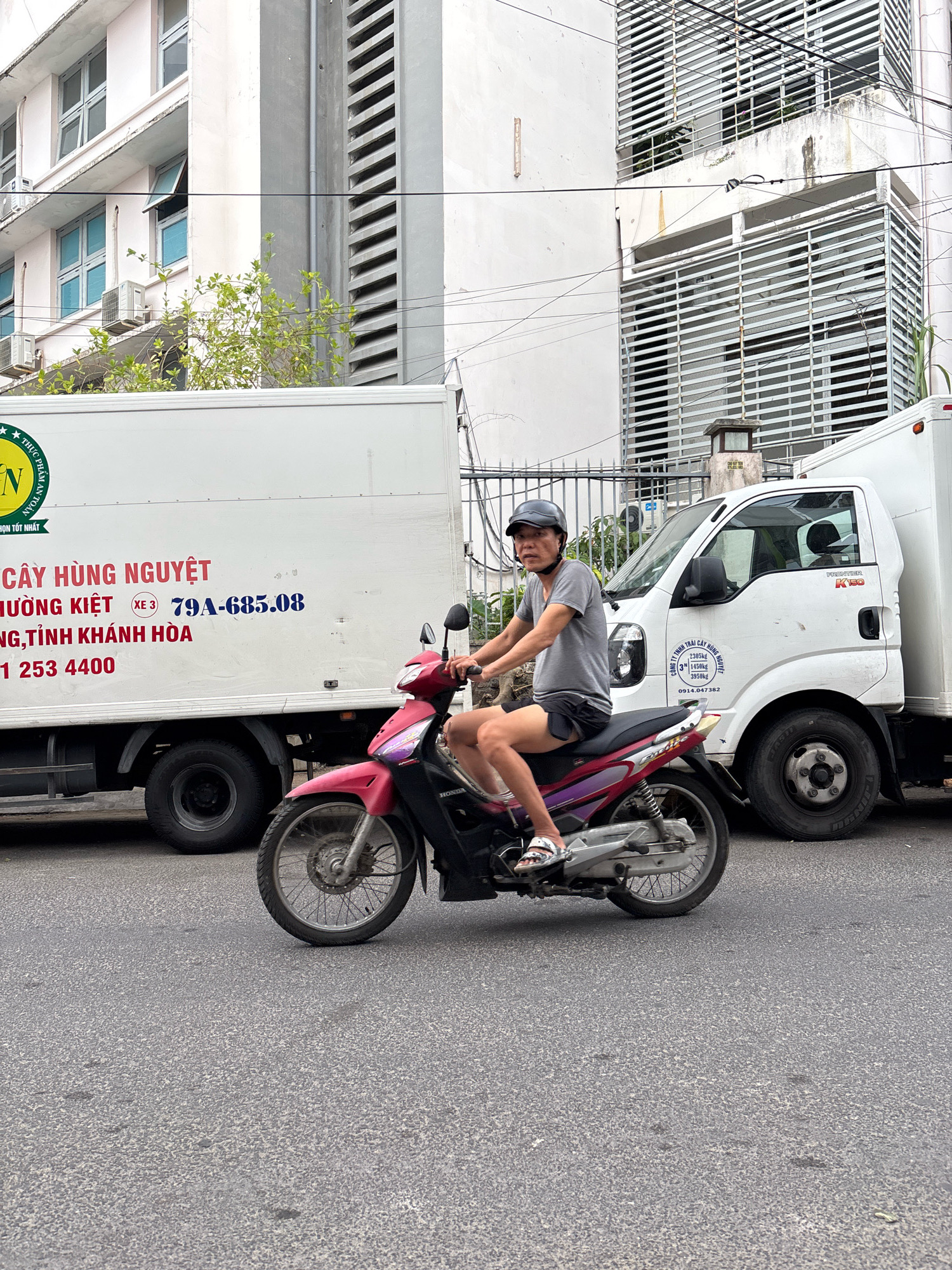 In Nha Trang, Vietnam, a man wearing a grey t-shirt, black shorts, a grey helmet, and white sandals rides a pink and black scooter along an asphalt road. He is positioned on the right side of the frame, facing left. Behind him, a white box truck with blue and red lettering is parked, and a portion of a white delivery truck is visible to the right. The background features a multi-story building with white walls, numerous windows, and horizontal louvers. Power lines crisscross the upper portion of the image, and some green foliage is visible between the building and the box truck. The overall scene depicts a typical street view in a Vietnamese city.