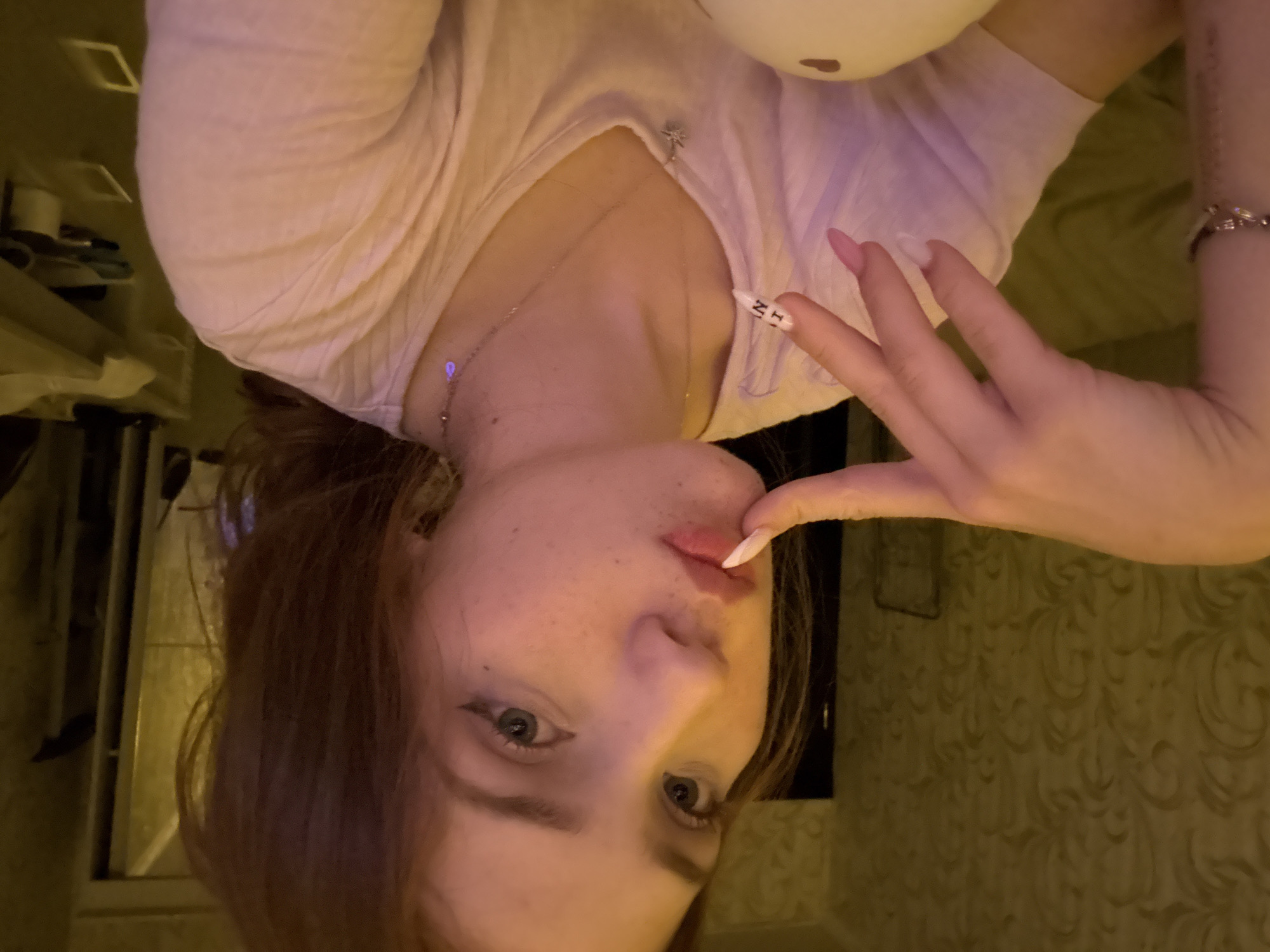 The image depicts a young woman in an indoor setting. She has light skin with visible freckles on her nose and cheeks, brown or auburn hair, and blue eyes with winged eyeliner. Her lips are lightly colored pink and slightly parted. She is wearing a light-colored, possibly pale pink or white, v-neck t-shirt. Her left hand is raised, with the index finger gently touching her lower lip. Her nails are long and manicured, primarily light pink, with the index finger nail being white and featuring indistinct black text (appearing as 'NIL' or 'LIN'). She wears a silver-toned bracelet on her left wrist and two delicate necklaces: one with a small star pendant and another with a small, iridescent teardrop charm. In the lower foreground, partially obscured by her body, is a large, smooth, light-colored, possibly white, rounded object. The background reveals an indoor environment. To the left, a wall is covered in patterned wallpaper with a swirling or floral motif in a muted golden-olive tone. To the right, parts of a room are visible, including a dark vertical element (possibly a door frame or furniture edge) and blurred light-colored furniture further back, with some indistinct items on a surface. The lighting is warm and artificial, casting soft illumination. There is no visual information within the image to specify the location as Naberezhnye Chelny, Russia, or any other specific city or country.