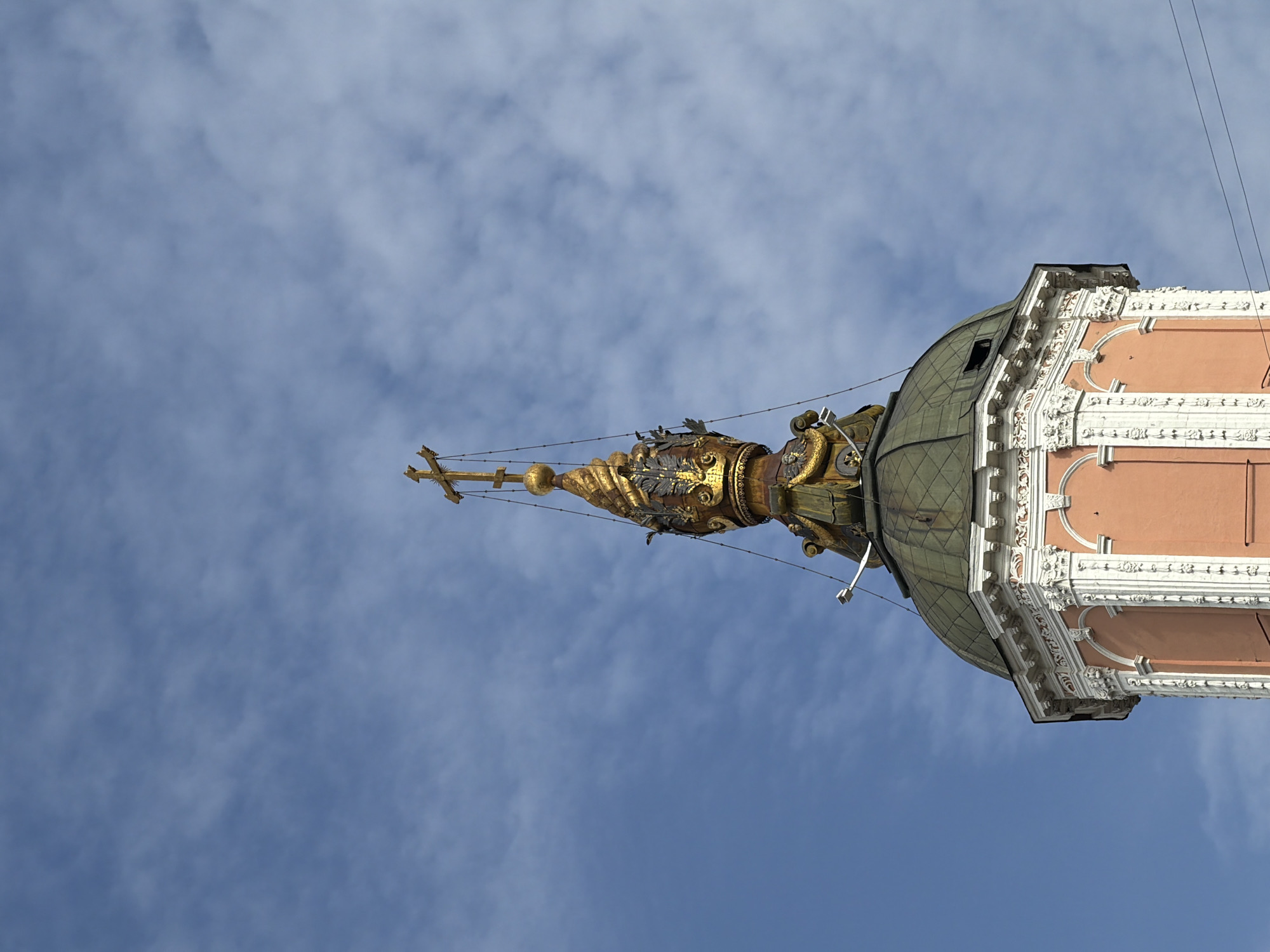 A low-angle shot of the top of a building with a golden spire topped by a cross, set against a blue sky with sparse clouds. The architectural style includes a dome with a green roof, an ornately decorated peach-colored facade with white trim, and gilded elements near the top of the structure. Thin cables are visible against the sky. The location is Moscow, Russia.