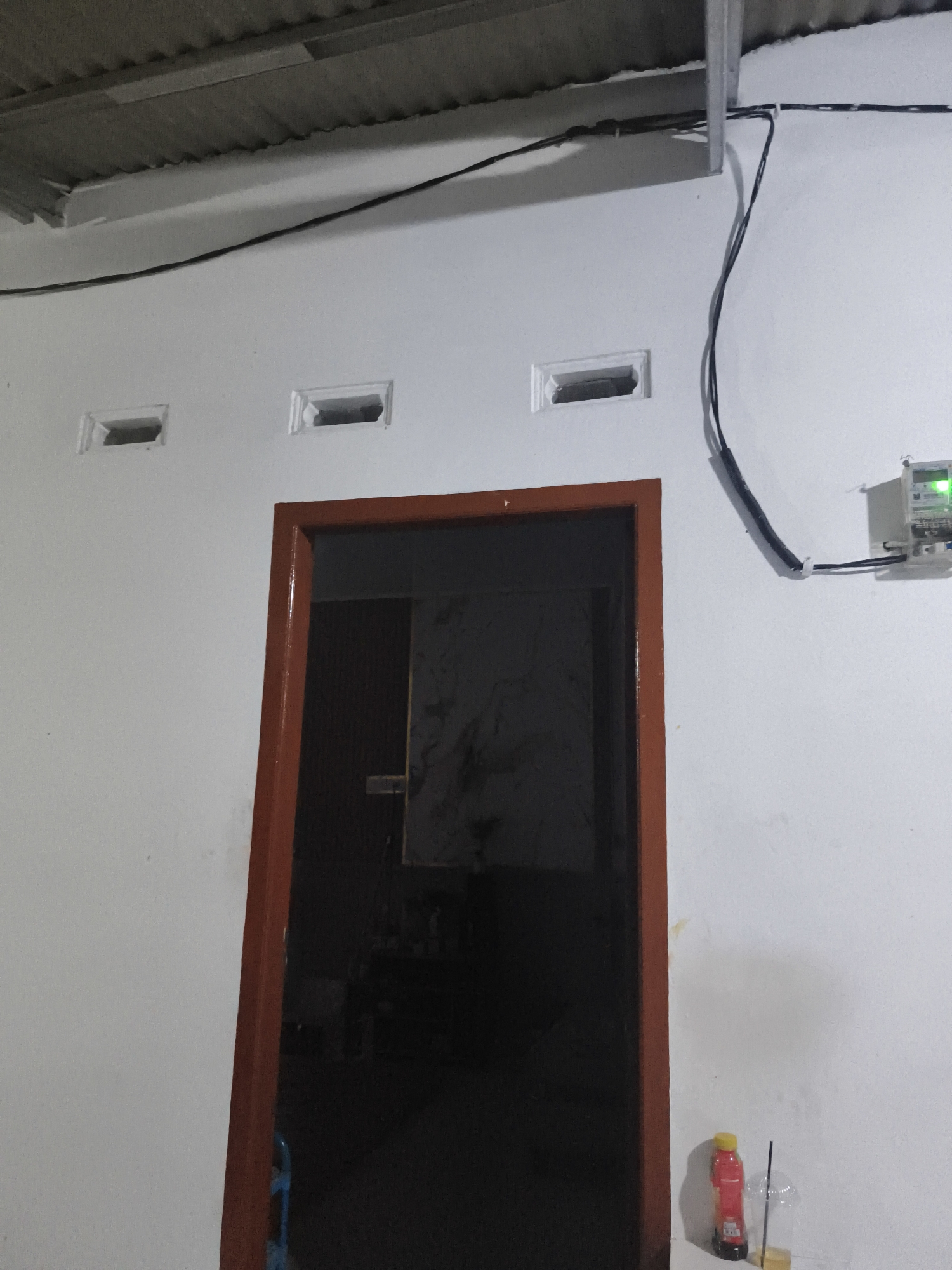 The image depicts the exterior of a building in Pangkalpinang, Indonesia, likely a residential home, showcasing a white wall, a dark doorway with a reddish-brown frame, and two small ventilation openings above the door. Electrical wiring runs across the wall, connecting to a visible electric meter box. In the lower right corner, a bottle of red liquid with a yellow cap and a clear plastic cup with a straw are placed on the ground. The scene appears to be captured at night or in low light conditions, judging by the general darkness and the illuminated electric meter.
