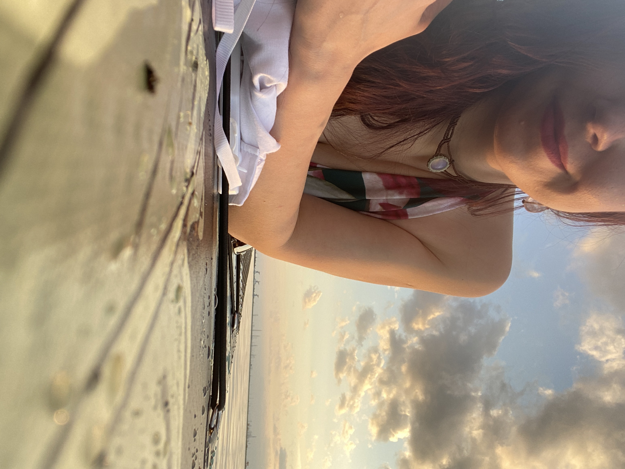 A woman with auburn hair is resting her arm on a wooden surface that appears to be part of a dock or deck. She is wearing a patterned top and a necklace with a pendant. The background features a body of water, possibly a bay or river, with the skyline of what seems to be Chaikovsky, Russia, visible in the distance. The sky is filled with clouds, suggesting either sunrise or sunset.