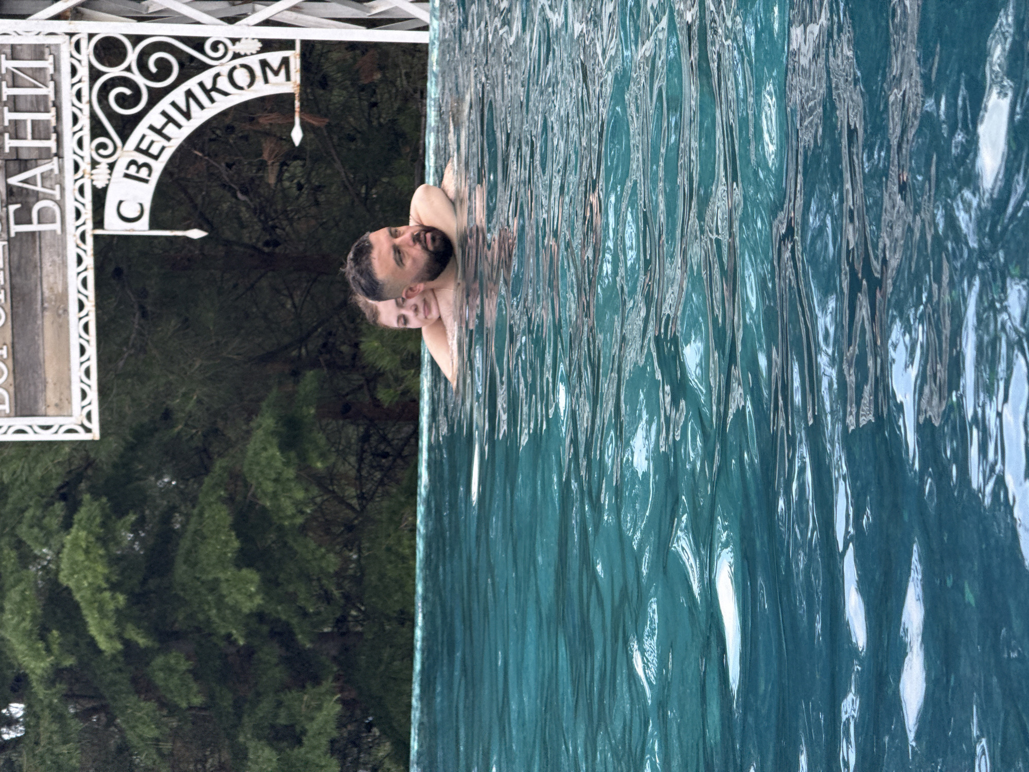In the city of Yalta, Ukraine, two people are in a swimming pool. A man with a beard is in the foreground, with a woman embracing him from behind, resting her head on his shoulder. The water is a vibrant turquoise color with visible ripples. In the background, lush green trees are visible, and a decorative sign with Cyrillic text is mounted on a white structure. The sign appears to read "БАНИ С ВЕНИКОМ," which translates to "Baths with a broom" or "Baths with a venik." The overall scene suggests a serene outdoor setting.