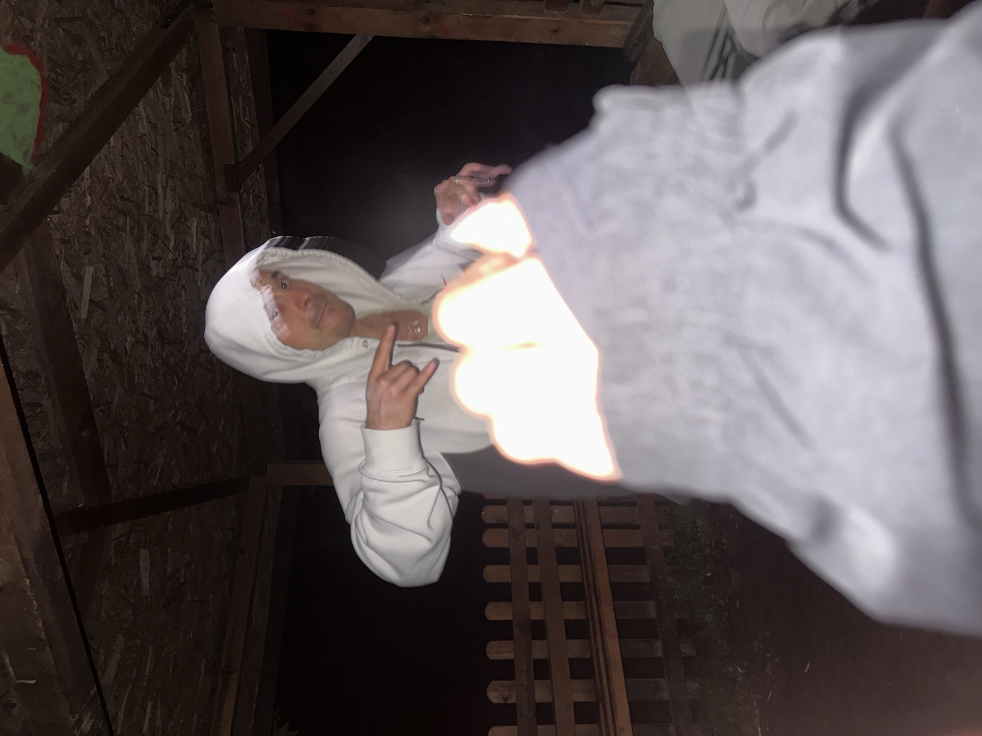 The image, rotated 90 degrees clockwise from its natural orientation, captures a person in a dimly lit, enclosed or semi-enclosed space. The central subject is an individual wearing a white hooded sweatshirt with the hood up, looking directly towards the camera with a neutral expression. Their left hand is raised, making a "rock on" or "devil horns" gesture.
The background features rough wooden planks and beams, suggesting an unfinished room, an attic, or a wooden shed. On the far left, an abstract shape in green and red is visible on a dark surface, possibly graffiti or a painted mark. The scene is predominantly dark, indicating a nighttime setting or an environment with minimal natural light.
A bright, out-of-focus light source, likely a camera flash, illuminates the right foreground, partially obscuring what appears to be a grey sleeve. Below this light and further in the background, a wooden lattice or fence structure is visible, hinting at an outdoor or semi-outdoor area adjacent to the main space. The overall mood is casual and informal. No discernible text is visible within the image. While the visual cues do not specify the exact location, the context suggests Blagoevgrad, Bulgaria.