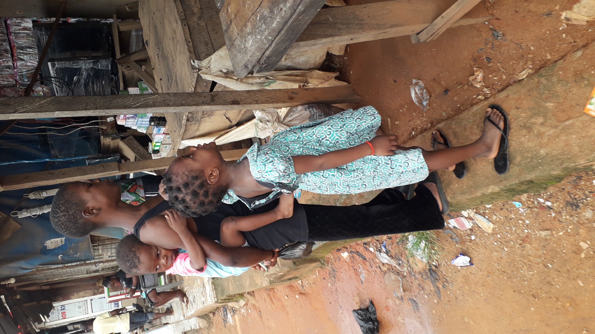 In Nkpor, Nigeria, a woman stands on a concrete curb, carrying a young child on her back. The child on her back is wearing a pink and blue shirt. Beside her, another young child in a teal and white patterned dress looks upwards. The background suggests a busy outdoor market or roadside stall area, with wooden structures, shelves stocked with various items, and a covered stall with a blue tarp. The ground is a mix of dirt and debris. The overall scene depicts a candid moment of everyday life in a Nigerian urban setting.