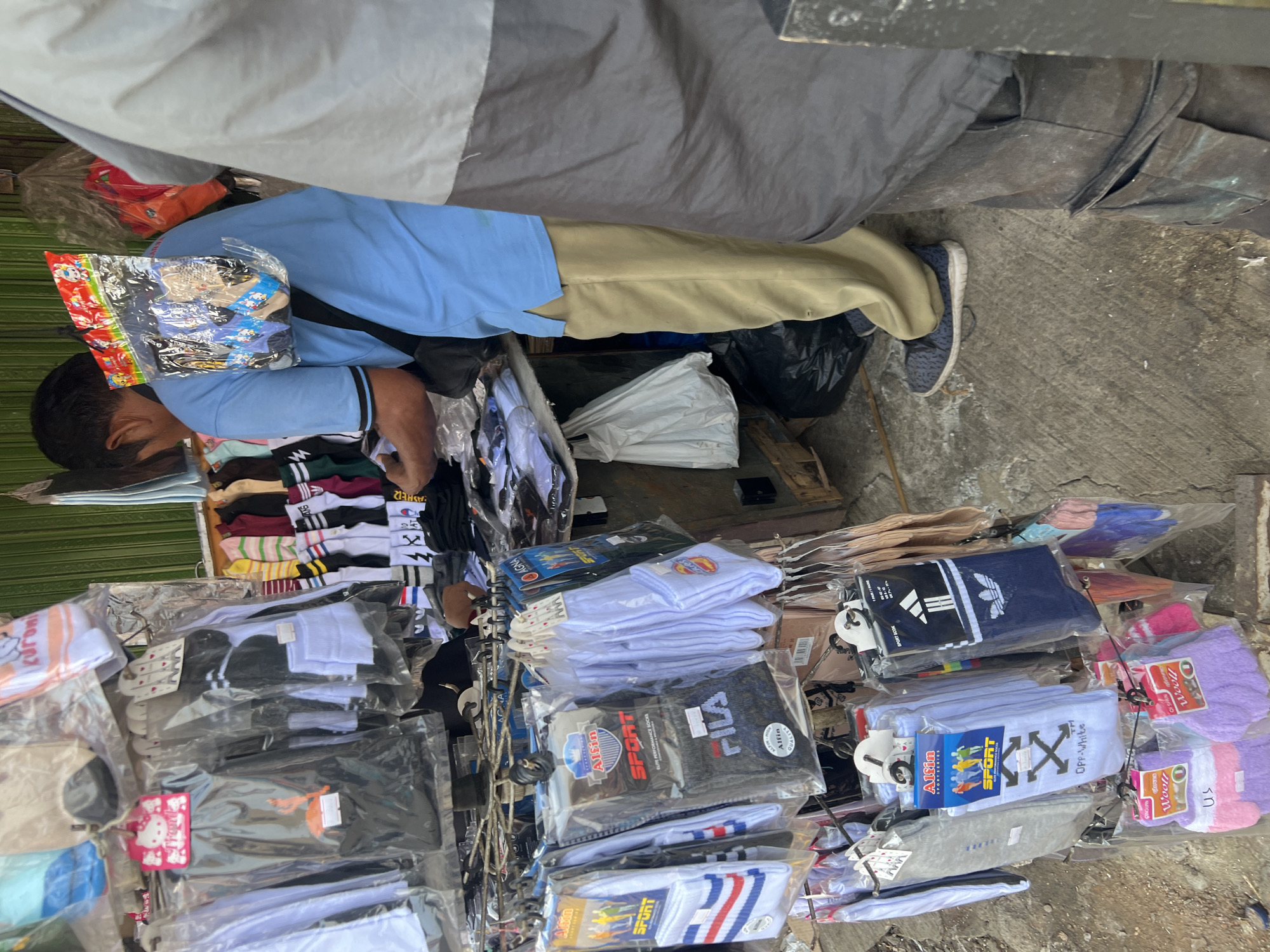 The image captures a street scene in Citeureup, Indonesia, likely a market stall. A man wearing a light blue polo shirt and tan pants stands in the center, with a package of socks attached to his back. He appears to be a vendor, as he is surrounded by racks of hanging socks and other clothing items, many of which are packaged. The stall itself is makeshift, with some items displayed on shelves and others hanging from a metal rack. In the background, a green corrugated metal wall is visible, suggesting an outdoor or semi-enclosed space. To the right of the central figure, another person, partially obscured by shadow and wearing a grey garment, stands close by. The ground is paved with concrete. The overall impression is one of a busy commercial environment, typical of a local market.
