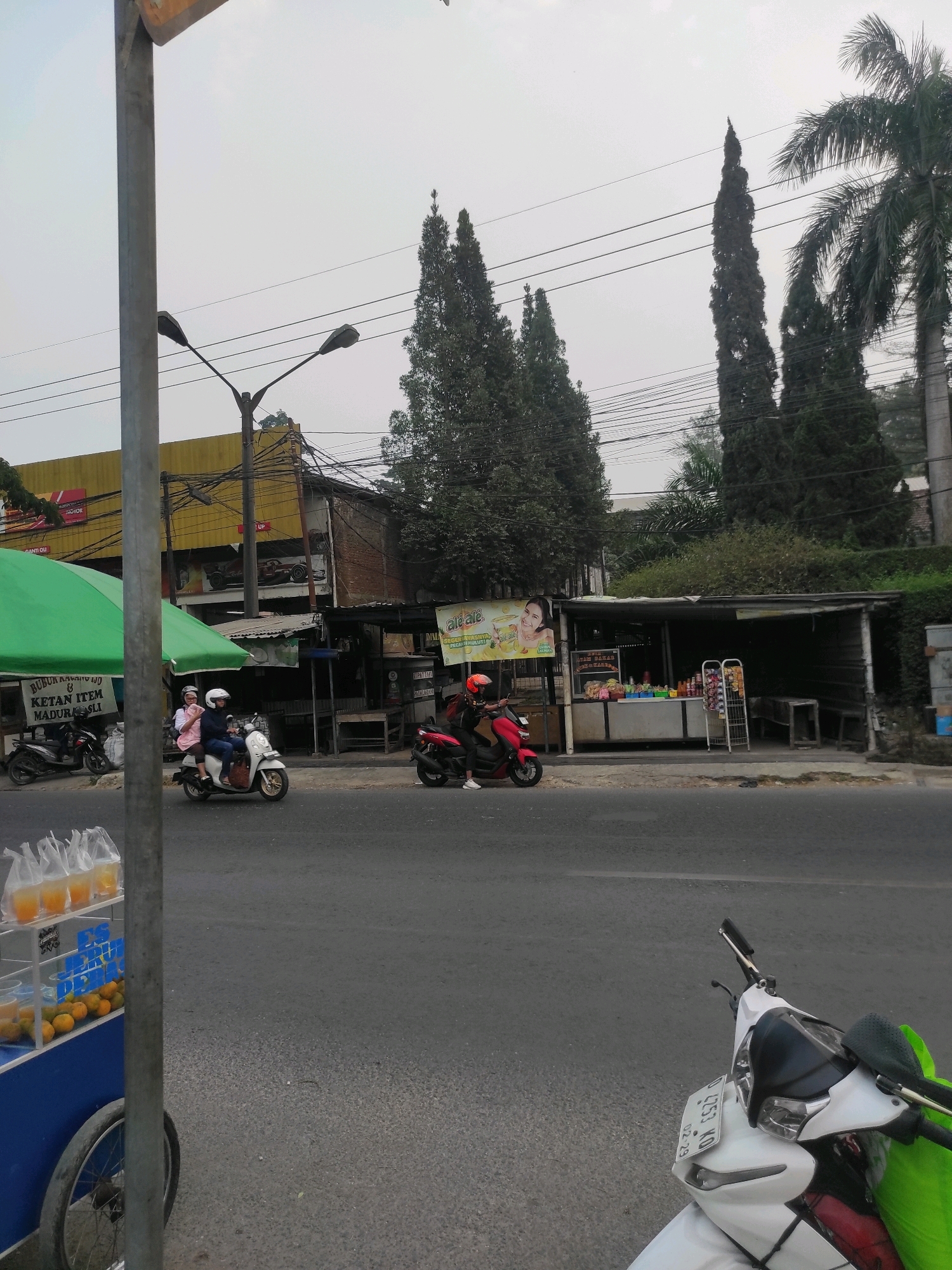 In the city of Ciburuy, Indonesia, a street scene unfolds under an overcast sky. A paved road occupies the foreground, with a white scooter parked on the right side. Further down the road, two individuals ride a white scooter. A vendor cart filled with oranges and bagged juices sits on the left, partially obscuring a green canopy. Behind the cart, a building with a yellow facade is visible, featuring a sign with red lettering. Adjacent to it, a small shop with an open front displays various products on shelves. Several trees, including tall cypress-like trees and a palm tree, line the background, with power lines strung between them. A few other motorcycles are parked in front of the shops.