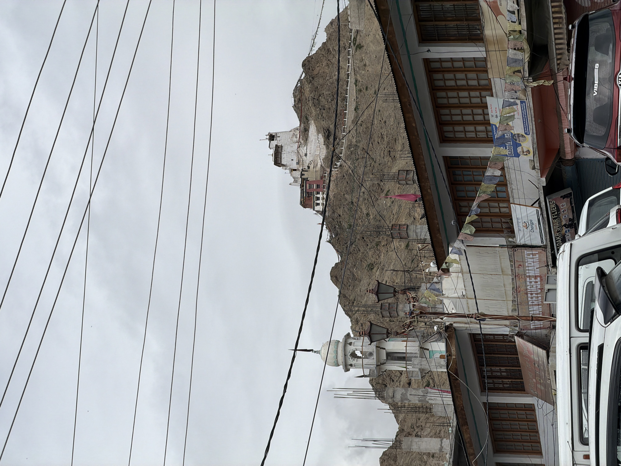 The image depicts a cloudy day in the city of Leh, India, with a view of buildings, a mosque, and a monastery perched on a rocky hillside. In the foreground, several white vans and a red SUV are parked on a street. Above the street, buildings with wooden-framed windows are visible, adorned with prayer flags. To the left, a white mosque with a dome stands tall against the mountainous backdrop. Further up the hill, the Leh Palace, a historic 17th-century palace and monastery complex, is prominently featured. The sky is overcast, suggesting a diffused light and potentially cool weather. The overall scene captures the unique architectural blend and the dramatic natural landscape characteristic of Leh.