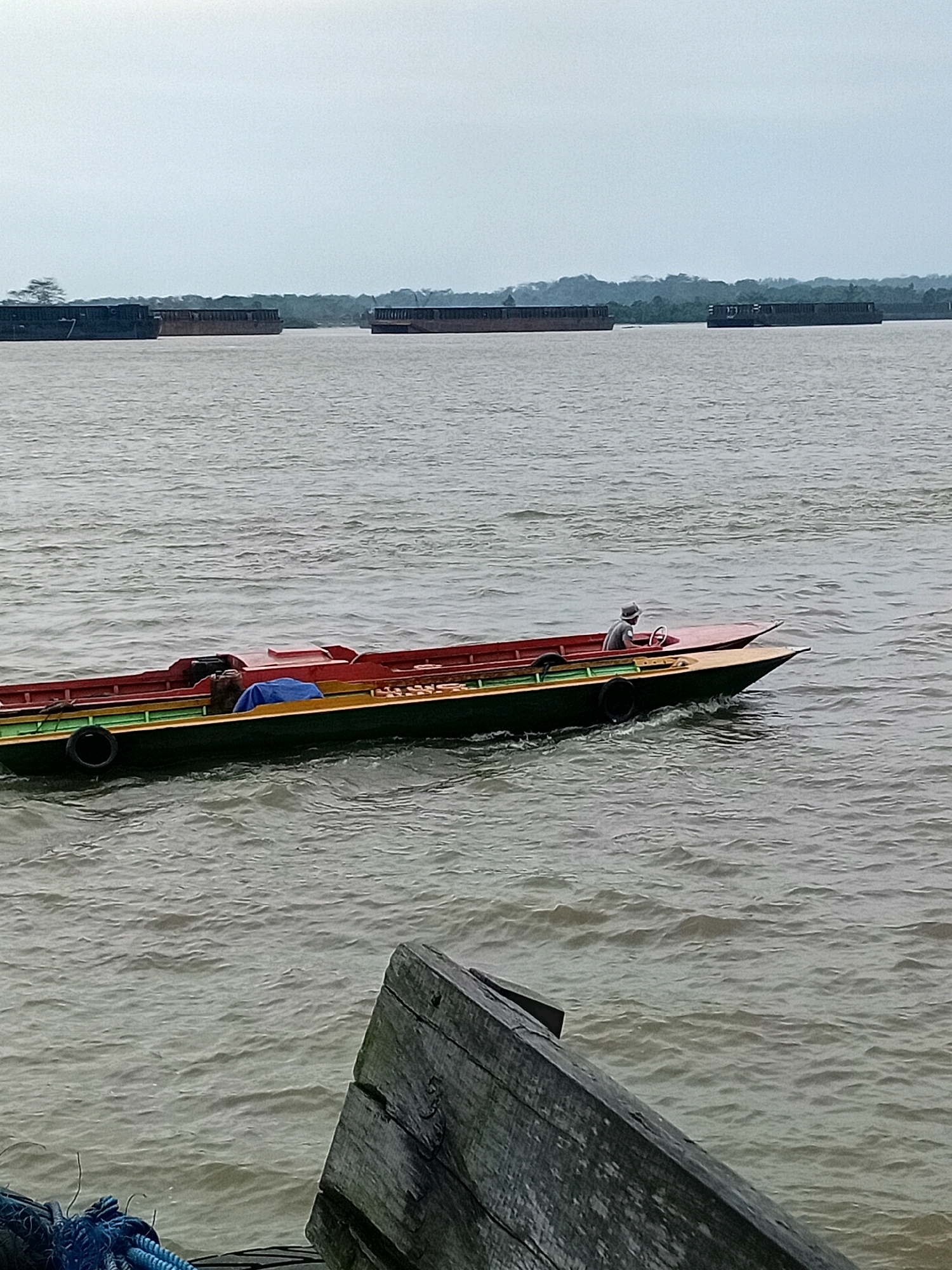 The image depicts a wide, murky river with several large barges in the background, some laden with cargo and others appearing empty. In the foreground, a small, narrow motorboat with a red and green hull is moving across the water. The boat is occupied by a single person wearing a hat, who is steering with a wheel. The boat also contains some cargo covered with a blue tarp, and two sleeping figures can be seen in the middle of the boat. The riverbanks in the distance are lined with dense green vegetation, suggesting a natural or semi-rural environment. In the lower foreground, a weathered, angled piece of wooden debris, possibly part of a dock or pier, is visible, with blue rope coiled near it. The sky is overcast, contributing to the muted light of the scene. The location is identified as Sungai Meriam, Indonesia.