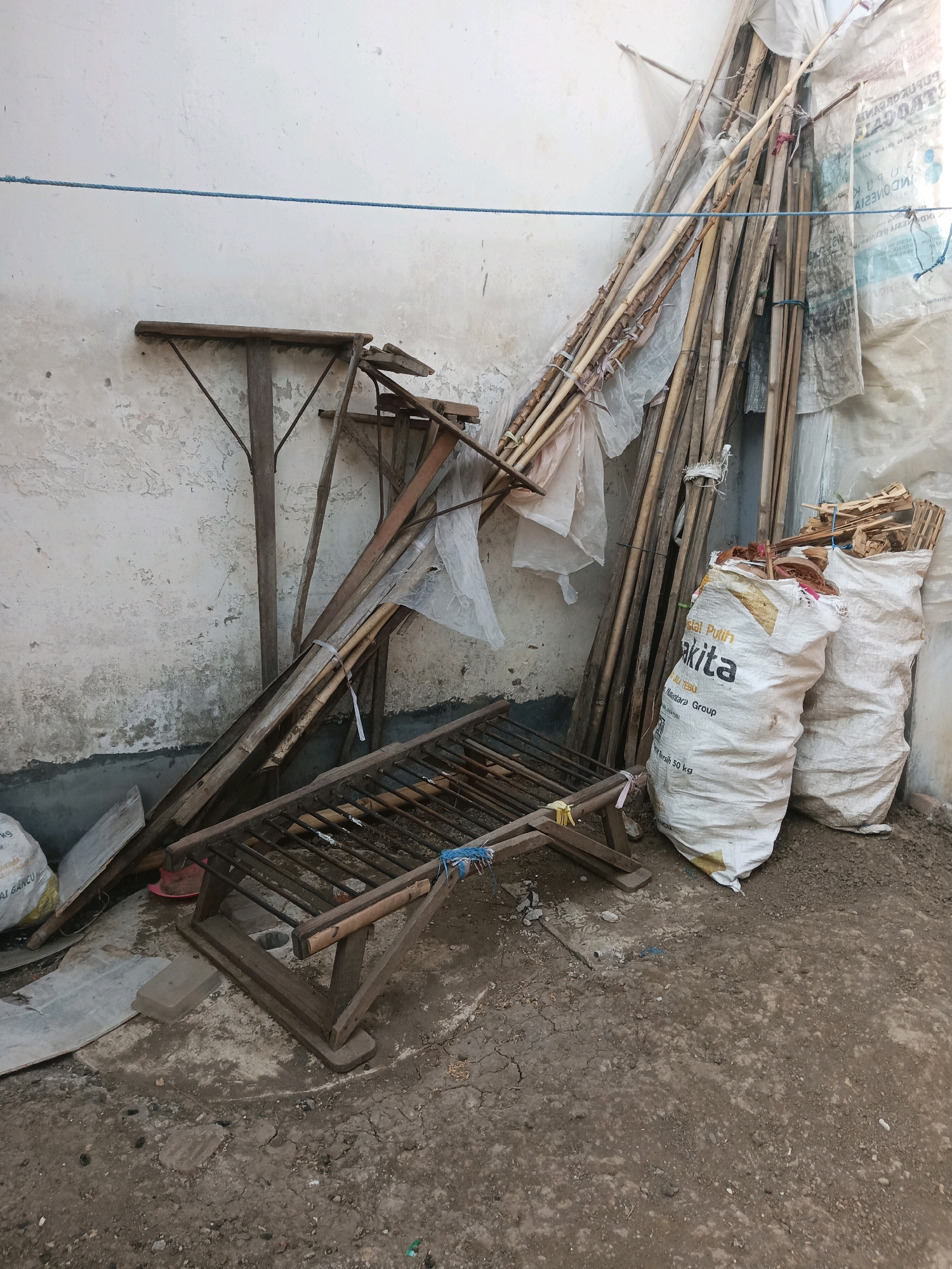 A vertical, eye-level, full shot shows a collection of stacked items against a textured white wall in an outdoor setting. The wall, with its rough and peeling surface, takes up the top half of the frame. A thin blue rope is stretched horizontally across the wall, approximately one-third of the way down from the top.
Leaning against the wall and extending into the foreground are various objects. A tall, leaning stack of long, thin bamboo poles, many wrapped with white plastic sheeting, dominates the right side of the image. Some of these poles appear weathered and tied together with string and white plastic ties. To the left of the bamboo, a dark wooden structure, possibly a disassembled table or stand with triangular supports, is propped up. Further to the left, a low, wooden, slatted frame rests on the ground. The frame has a dark metal grate or support structure visible within its wooden base.
In the foreground, two large, white woven sacks are visible. The sack on the left is partially obscured by the wooden structure. The sack on the right is more prominent and displays text in an orange and black font. Some of the text on the visible sack reads "TAKITTA" and "PT SUMBER." These sacks appear to be filled with a granular or loose material, possibly soil or fertilizer. Scattered on the ground around the sacks and the wooden structures are small pieces of wood and debris.
The ground is a mix of compacted dirt, loose soil, and small stones, with some areas appearing dry and cracked. The overall scene suggests a storage area or a yard where materials are being kept. The lighting is somewhat diffuse, indicating it might be daytime but possibly overcast or the area is in shadow. The environment appears to be an outdoor space, likely a yard or a storage area in a residential or semi-industrial setting in Ngrendeng, Indonesia. There are no people visible in the image.