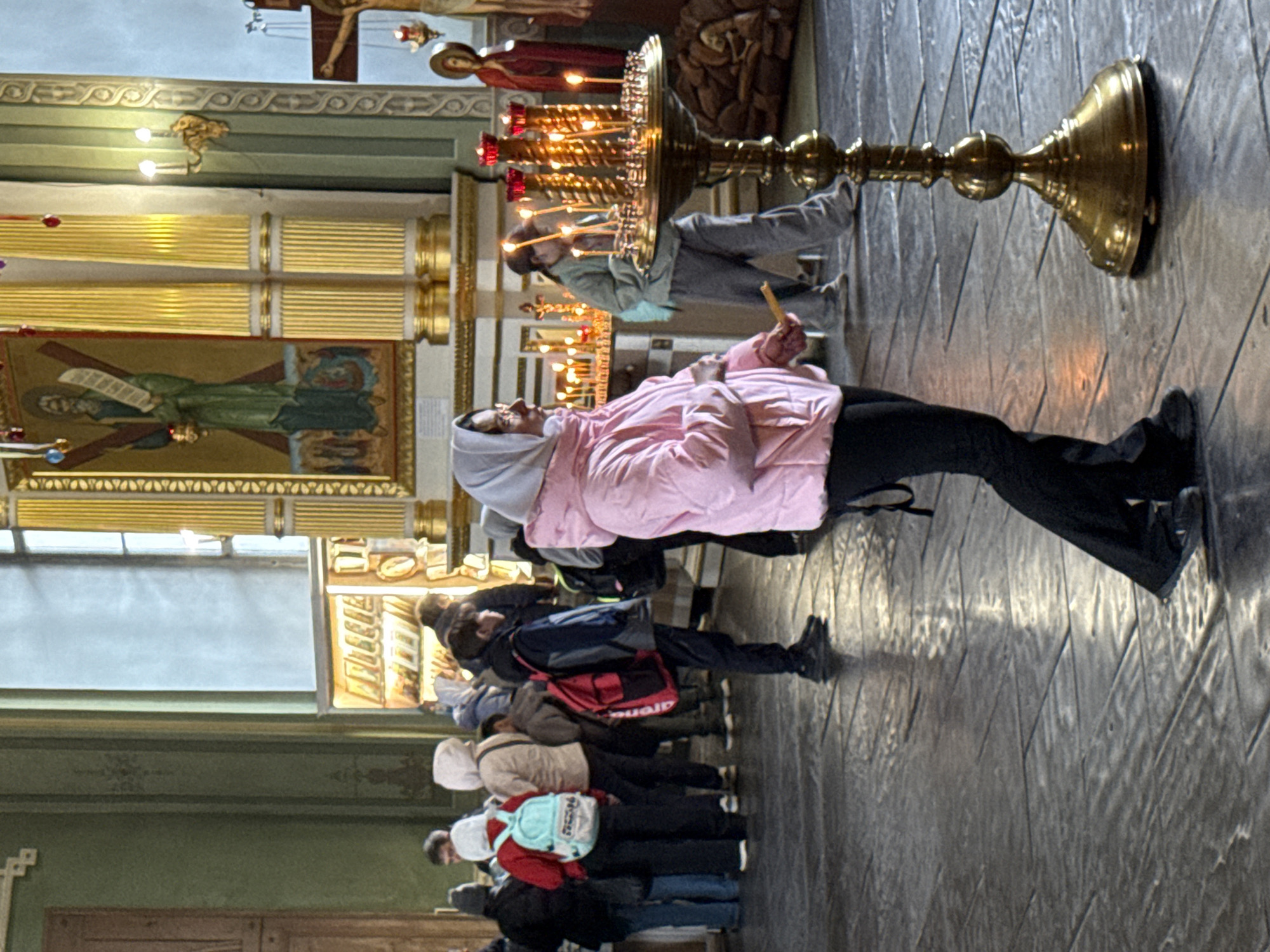 In a city of Kazan, Russia, in a church, a line of people standing in a queue is visible, each holding a candle. A woman in a pink jacket and head covering is placing a candle on a tall brass candlestick covered in lit candles. The church has green walls, gold accents, icons on the wall, and windows, with a cruciform structure at the top of the image. The floor is a diamond patterned wood or stone.