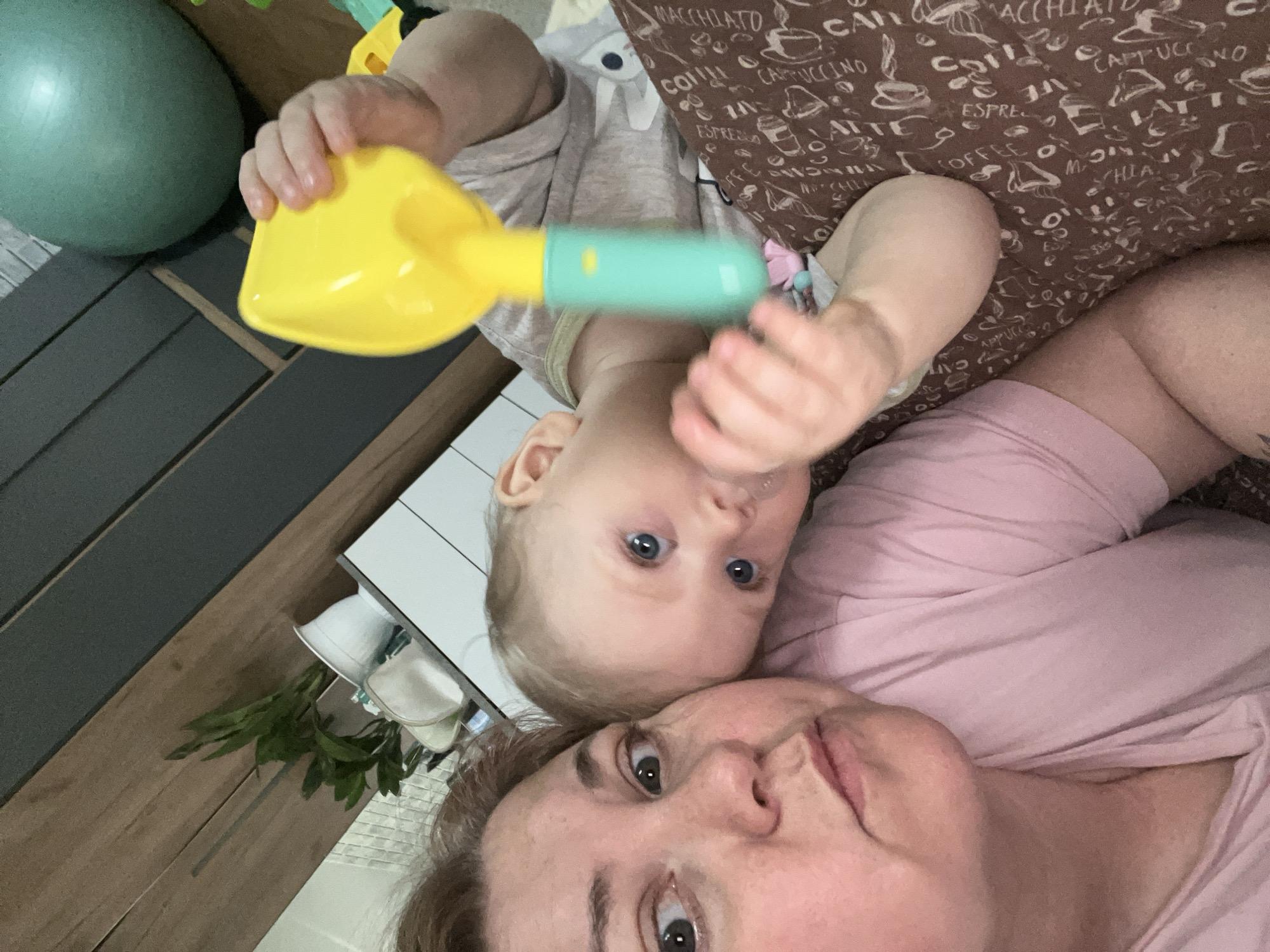 An indoor, close-up photograph features an adult woman and a baby lying down together, both looking directly at the camera. The woman, with brown hair and eyes, is in the lower half of the frame, wearing a light pink top. Above her, the baby, with light blonde hair and bright blue eyes, is positioned, holding a yellow and mint-green toy shovel or scoop.
The setting appears to be a casual home environment in Vesyoloye, Russia. Behind the baby, a brown patterned fabric or pillow is visible, adorned with various coffee-related words such as "MACCHIATO," "COFFEE," "CAPPUCCINO," "ESPRESSO," and "LATTE." To the upper left, a large, greenish-blue exercise ball rests on a dark wooden surface, which continues across the middle left of the frame. A small green leafy plant is partially visible near the bottom left edge, alongside a white surface. The scene captures an intimate moment, likely between a mother and child, with soft, natural lighting suggesting it is daytime.