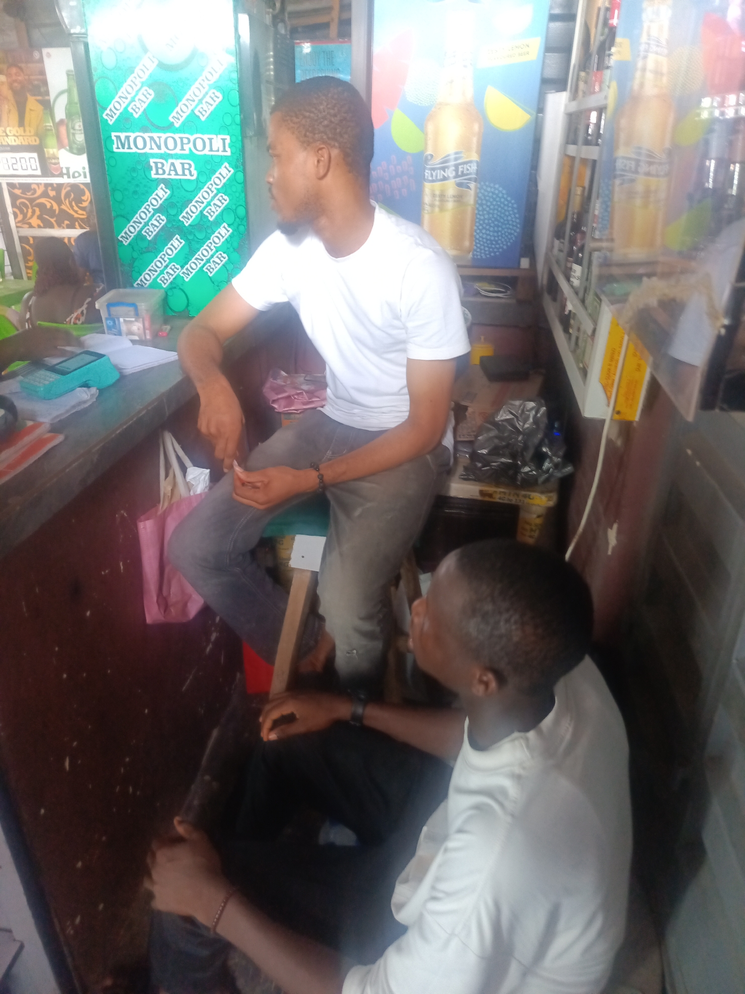 The image depicts the interior of a small establishment, possibly a bar or convenience store. Two young Black men are present. The man on the left is seated, wearing a white t-shirt and grey jeans, and appears to be interacting with a cash register or calculator on a counter. A pink tote bag hangs from a stool next to him. Behind him, a green cooler with "MONOPOLI BAR" branding is visible, along with other advertisements for drinks, including "FLYING FISH." The man on the right is also seated, facing away from the camera, wearing a white t-shirt and dark trousers. He is looking to his left, engaged in conversation with the other man. The environment suggests a casual, possibly dimly lit setting, with various items on shelves and counters, including bottles, bags, and other merchandise.