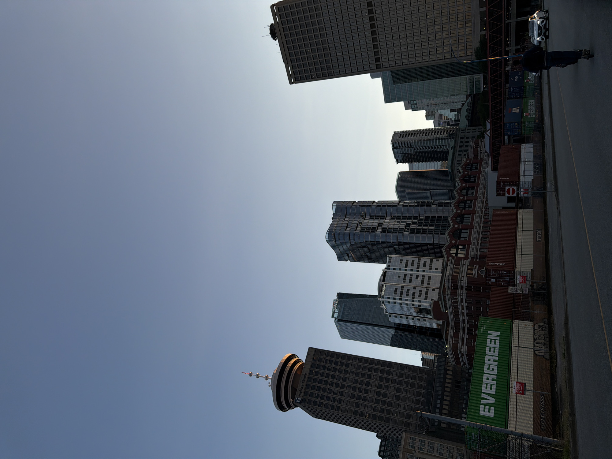 The image shows a city skyline in Vancouver, Canada, with a clear blue sky. In the foreground, there is a street with a person riding a scooter. To the left, several shipping containers are stacked, with one prominent green container labeled "EVERGREEN." In the background, numerous skyscrapers of varying heights and architectural styles dominate the view. Notably, the Harbour Centre tower, with its distinctive observation deck, is visible. The scene appears to be during the golden hour, with the sun casting long shadows and illuminating the buildings. The overall atmosphere is one of an urban environment, possibly during the late afternoon or early evening.