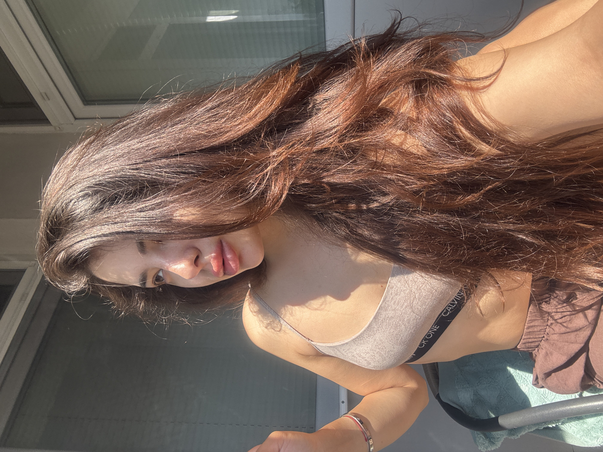 A woman is depicted in an outdoor setting, presumably on a balcony in Plovdiv, Bulgaria, illuminated by strong, direct sunlight that casts prominent highlights and shadows. She is presented in a profile view, looking towards the left side of the frame. Her long, wavy, dark brown hair, exhibiting distinct auburn tones under the sunlight, flows over her right shoulder and chest. Her complexion is fair to lightly tanned, with bright highlights visible on her cheekbone and the bridge of her nose. She is attired in a light-colored (beige or light grey) bralette or sports bra, the elastic band of which prominently displays the text "CALVIN KLEIN CK ONE". Below this, she wears loose-fitting, brown or rust-colored pants or shorts. A thin bracelet, featuring red and silver elements, is visible on her left wrist. She appears to be seated, with a portion of a chair covered by a teal or light blue fabric visible on the left edge of the frame. The background consists of a building's exterior, including windows with discernible horizontal blinds or shutters visible through the glass, and white window frames. The scene is brightly lit, indicative of a clear, sunny day.