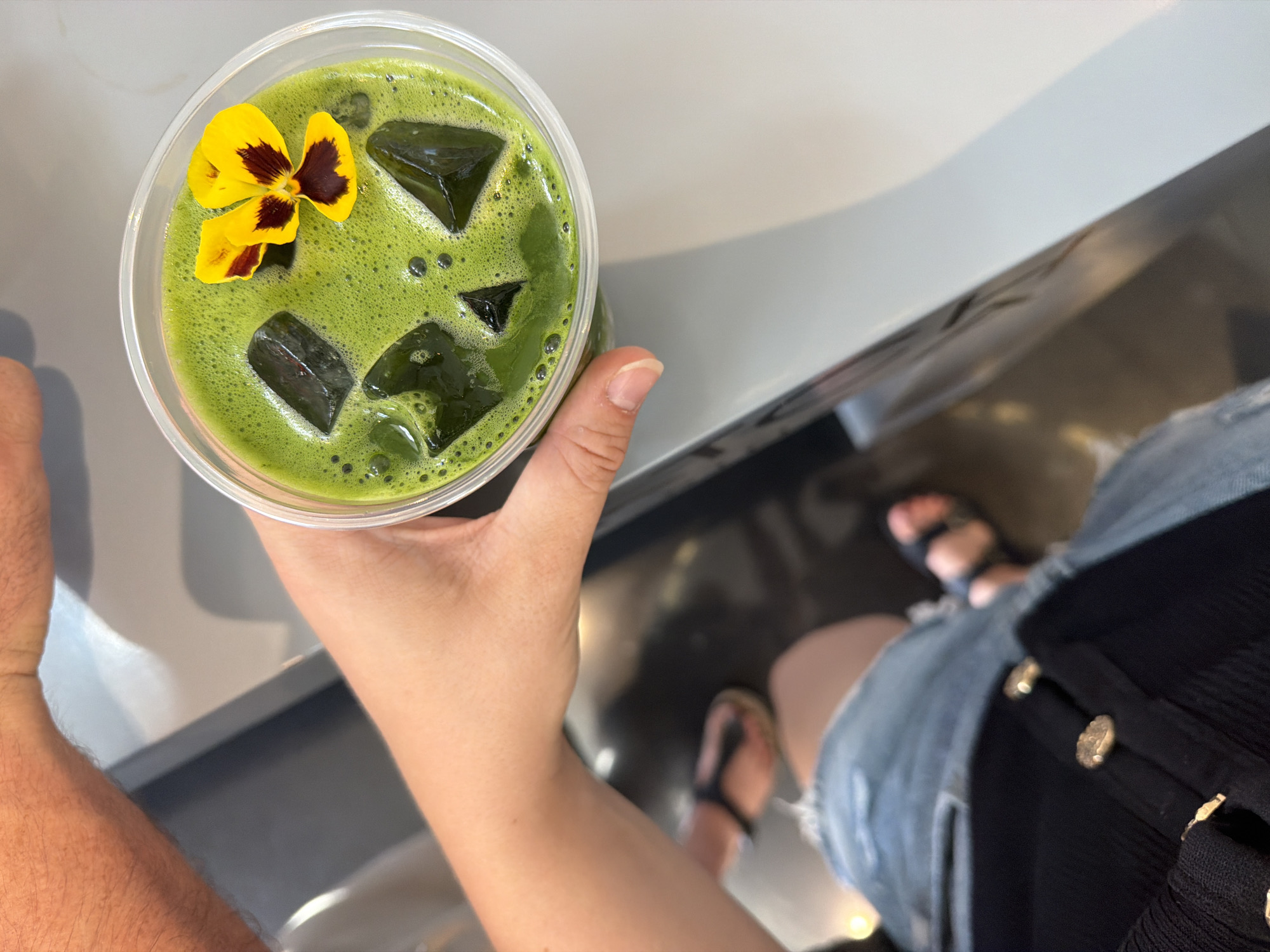 In a top-down perspective, a person's hand holds a clear plastic cup filled with a vibrant green, frothy beverage. Ice cubes are visible within the drink, and a yellow and purple pansy is delicately placed on the surface. The background is a blurred, neutral-toned interior, suggesting a cafe or restaurant setting, possibly in Munich, Germany. The person is wearing ripped denim shorts and a black top with gold buttons. The presence of other hands in the frame indicates multiple people are present.