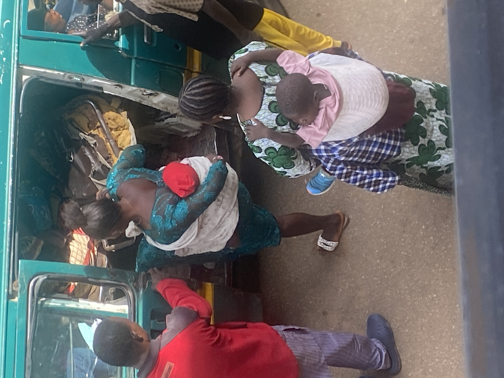 In Bukuru, Nigeria, a scene unfolds involving several individuals and a damaged vehicle. The primary focus is a green van, its door open and interior appearing disheveled with visible damage to seats and debris.
Several people are engaged in activities around the van. In the foreground, a person wearing a red long-sleeved shirt and grey striped trousers stands with their back to the camera, looking towards the van. To the right, a woman dressed in a vibrant green and white patterned dress, her hair in braids, is seen from behind, carrying a child on her back. This child is wrapped in a pink and white cloth. Another woman, wearing a teal and blue patterned garment, is also carrying a child on her back, this child is wrapped in white and has a red object near its head. The woman in teal is also wearing a white cloth around her shoulders.
Further to the right, a person in a patterned shirt and yellow trousers is partially visible, facing away from the camera and toward the interior of the van. Inside the van, a glimpse of another person can be seen through the side window, wearing a black and white patterned headscarf.
The ground is a rough, textured surface, likely a road or pathway. The overall impression is one of activity and perhaps a situation requiring assistance or attention, given the state of the vehicle and the engagement of multiple people.