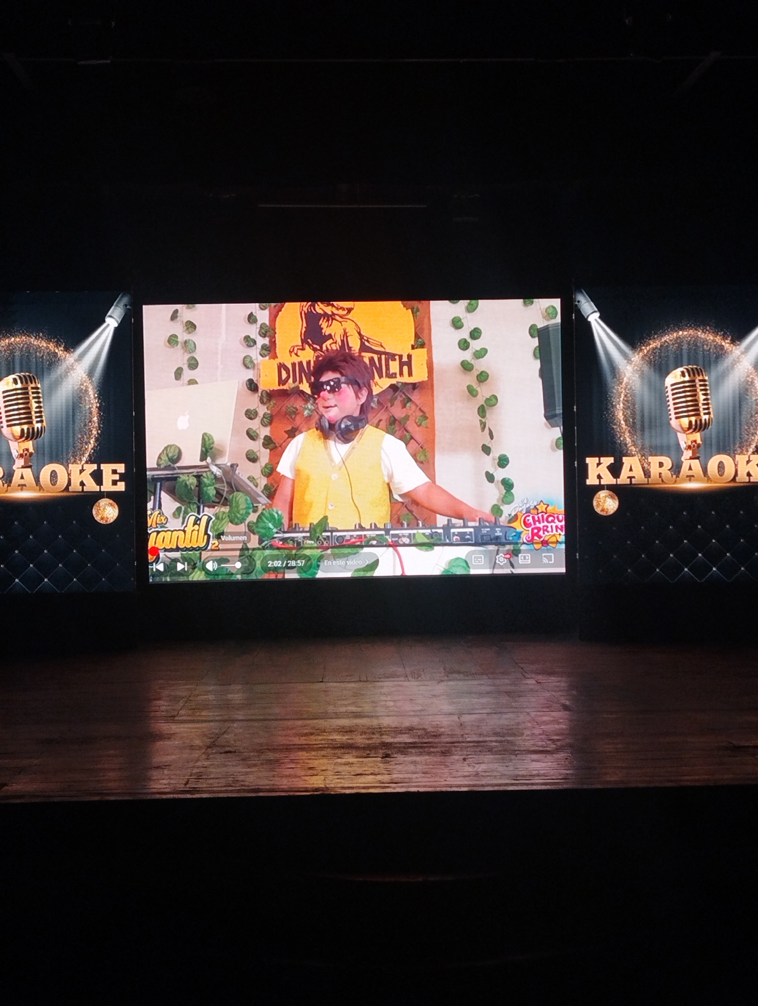 The photograph captures a stage setup, likely for karaoke entertainment. In the center is a large screen displaying a video featuring a person wearing sunglasses and headphones and a yellow vest, operating what appears to be a DJ console. Behind this person is a backdrop with vegetation and a sign. The video on the screen seems to be playing from a platform like YouTube, with playback controls visible at the bottom. Flanking the central screen on either side are smaller screens displaying a graphic design of a vintage microphone with the word "KARAOKE" written beside it. The screens are illuminated and contrast with the darker environment. The stage floor is wooden and dimly lit, with the darkness extending into the foreground. The scene gives the impression of a karaoke event or show being recorded or projected. It's not possible to ascertain its location in Porlamar, Venezuela from the content of the image.