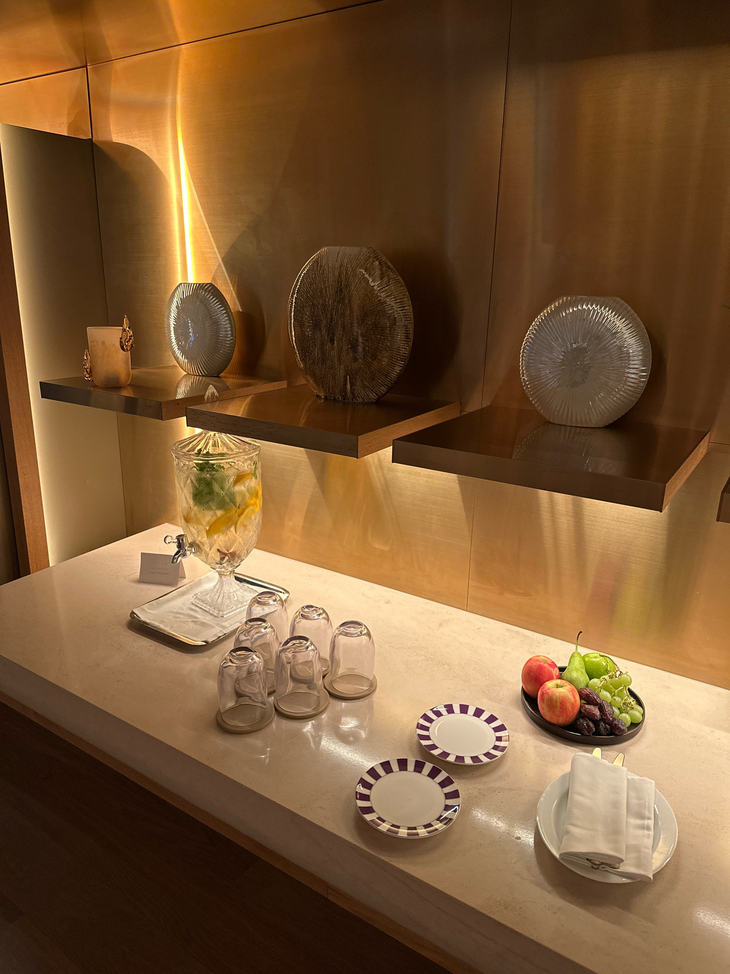 The image depicts a beverage station set up in what appears to be a spa or hotel lobby in London, United Kingdom. On a light-colored countertop, a clear glass beverage dispenser filled with water, lemon slices, and mint leaves is prominent. Beside it, several empty glasses are arranged upside down. A small silver tray holds a card that reads "THE SPA." To the right, a black platter contains a selection of fruits including red apples, green pears, green grapes, and dates. Two small plates with purple and white stripes are also visible, along with a white plate holding folded white napkins and cutlery. In the background, two wooden shelves are mounted on a brushed gold wall, decorated with three decorative glass or ceramic objects. The lighting is warm and ambient, highlighting the textures of the wall and the objects. There are no people present in the image.