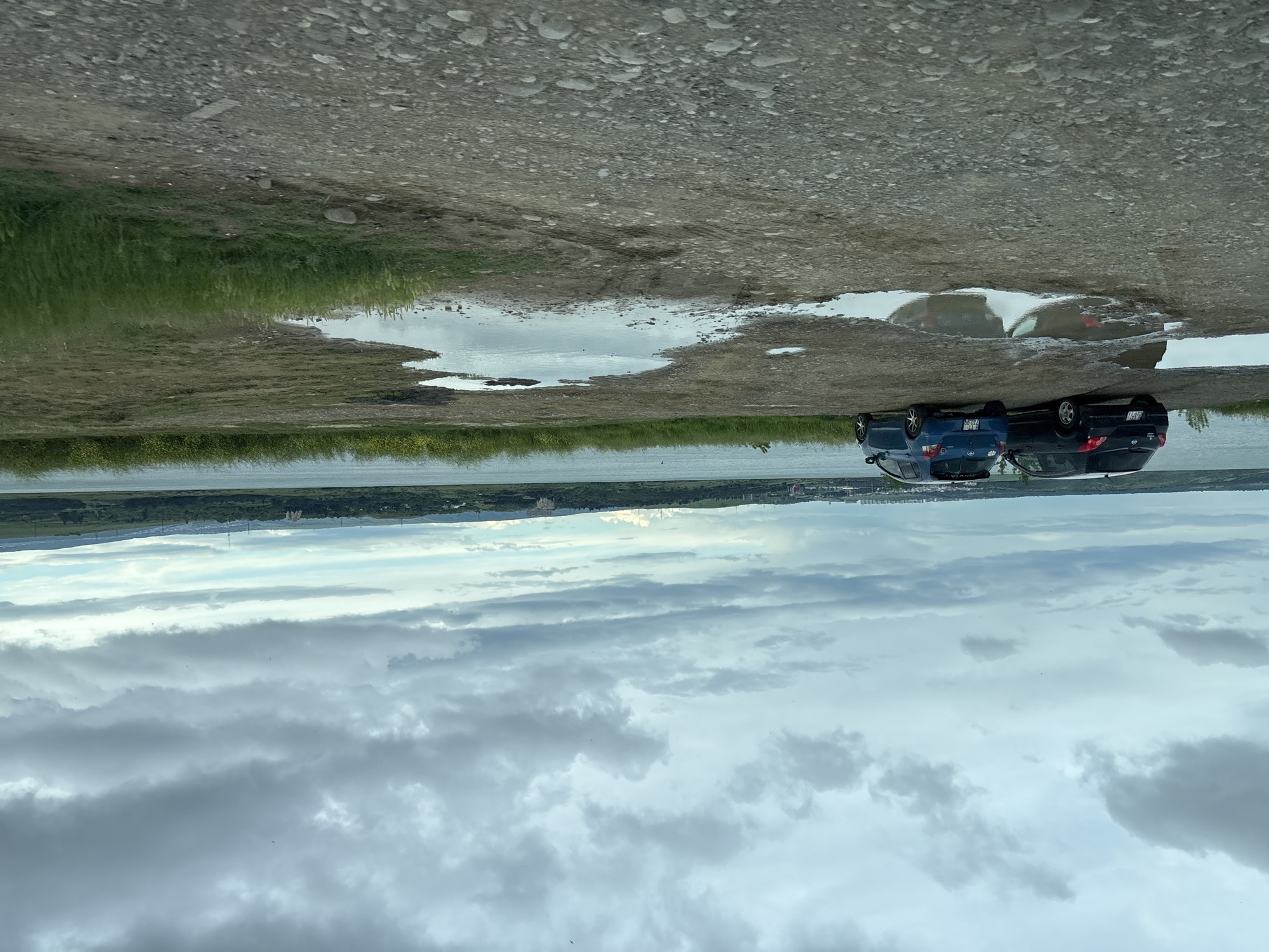 The image depicts a roadside scene with two cars parked near a body of water, likely near Tbilisi, Georgia. The background consists of a distant skyline, partially obscured by clouds. The foreground features a dirt road with puddles of water reflecting the sky and the parked cars. The sky is filled with gray, overcast clouds. Two compact cars, one blue and one black, are parked side-by-side on the edge of the road, near the water's edge. The environment is a mix of natural and developed landscapes. The cars appear to be parked near the Tbilisi Sea.