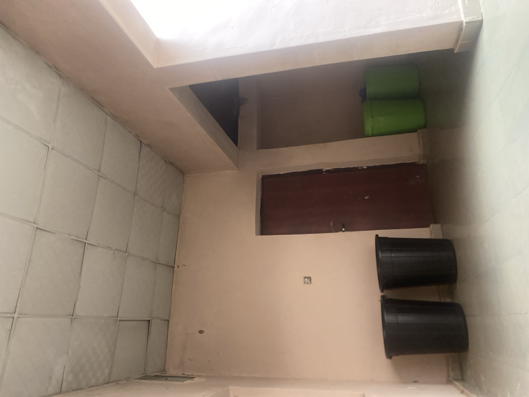 The image depicts an interior scene, likely a room or hallway in Wauro Leggal, Nigeria. The walls are painted in neutral tones, with a section of tiled wall on the left. Above, a bright light source suggests daytime or a well-lit interior space.
Prominent objects in the scene include two large, black cylindrical bins positioned vertically on the floor. In an elevated alcove, two bright green plastic containers are stacked. A dark wooden door or panel is visible on the wall, partially obscured by the alcove. There is a small, square electrical outlet on the peach-colored wall.
The scene appears to be a storage area or a utility space within a building. There are no people visible, and no specific activity is taking place. The setting is utilitarian, with an emphasis on practical storage solutions. The overall impression is one of a simple, functional interior. No text is visible in the image.