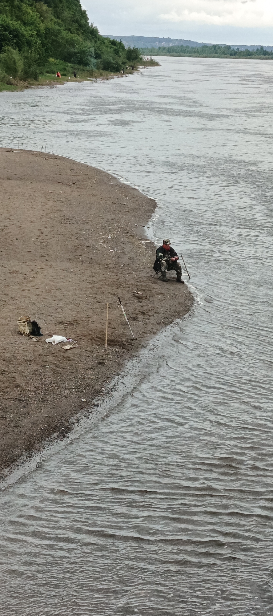 The image depicts a wide, grey-brown river bordered by a sandy/dirt bank and a steep, tree-covered hillside under an overcast sky, in what is identified as the city of Sarapul, Russia.
In the mid-ground, at the center-right, a person dressed in camouflage attire, including a cap and a red facial covering, is seated on a dark folding chair directly at the water's edge. This individual holds a fishing rod angled into the river. A thin wooden stick is positioned vertically near their right leg.
To the immediate left of the seated individual, on the sandy bank, several items are laid out: a light-colored fabric bag, a dark piece of cloth or bag, a white plastic bag or sheet, and a small, flat wooden object. Two sticks are stuck into the sand: a light wooden pole on the left and a thinner, dark-colored pole leaning to the right.
Further down the bank to the left, multiple fishing rods are visible, standing upright with their tips extending over the water. Several other individuals are dispersed along the riverbank in this area, also appearing to be engaged in fishing.
The river itself exhibits moderate ripples and small waves, suggesting a gentle current. Across the river in the far distance, low-lying land with sparse vegetation and faint outlines of structures is visible beneath the cloudy horizon. The hillside along the near bank is densely covered with green foliage. The lighting is diffuse, indicative of an overcast daytime.